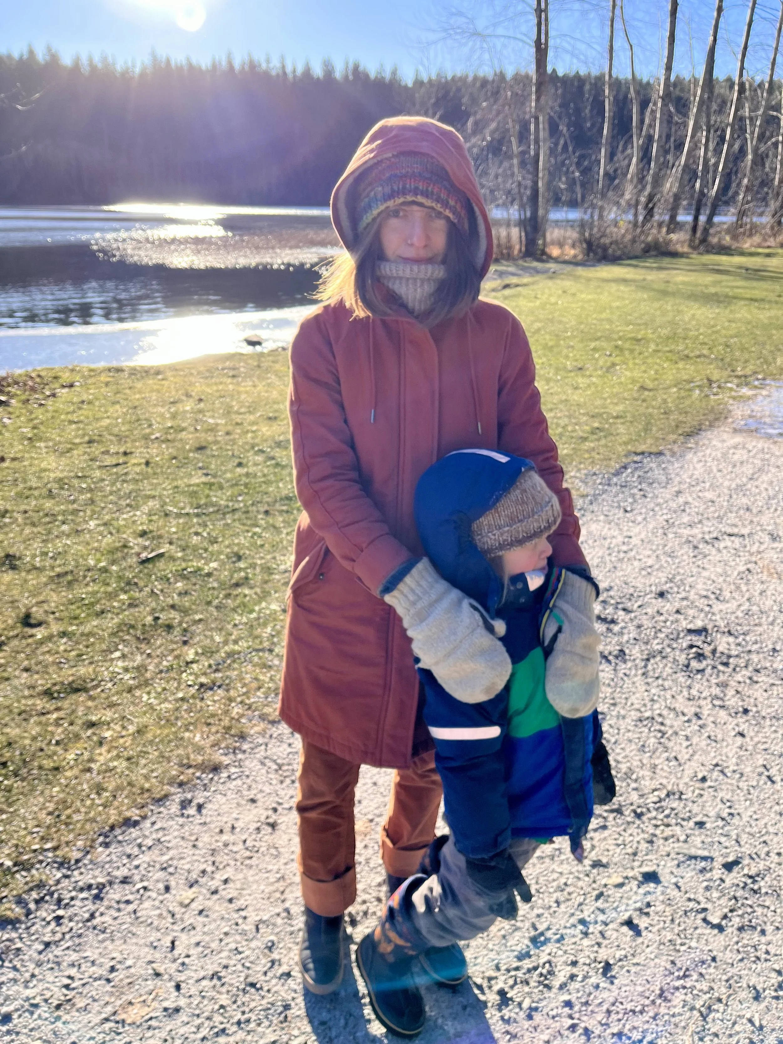 A woman and a young child outdoor near a lake in winter, both dressed warmly with jackets, hats, and gloves, standing on a gravel path with snow and grass around, with trees and water in the background and the sun shining brightly.