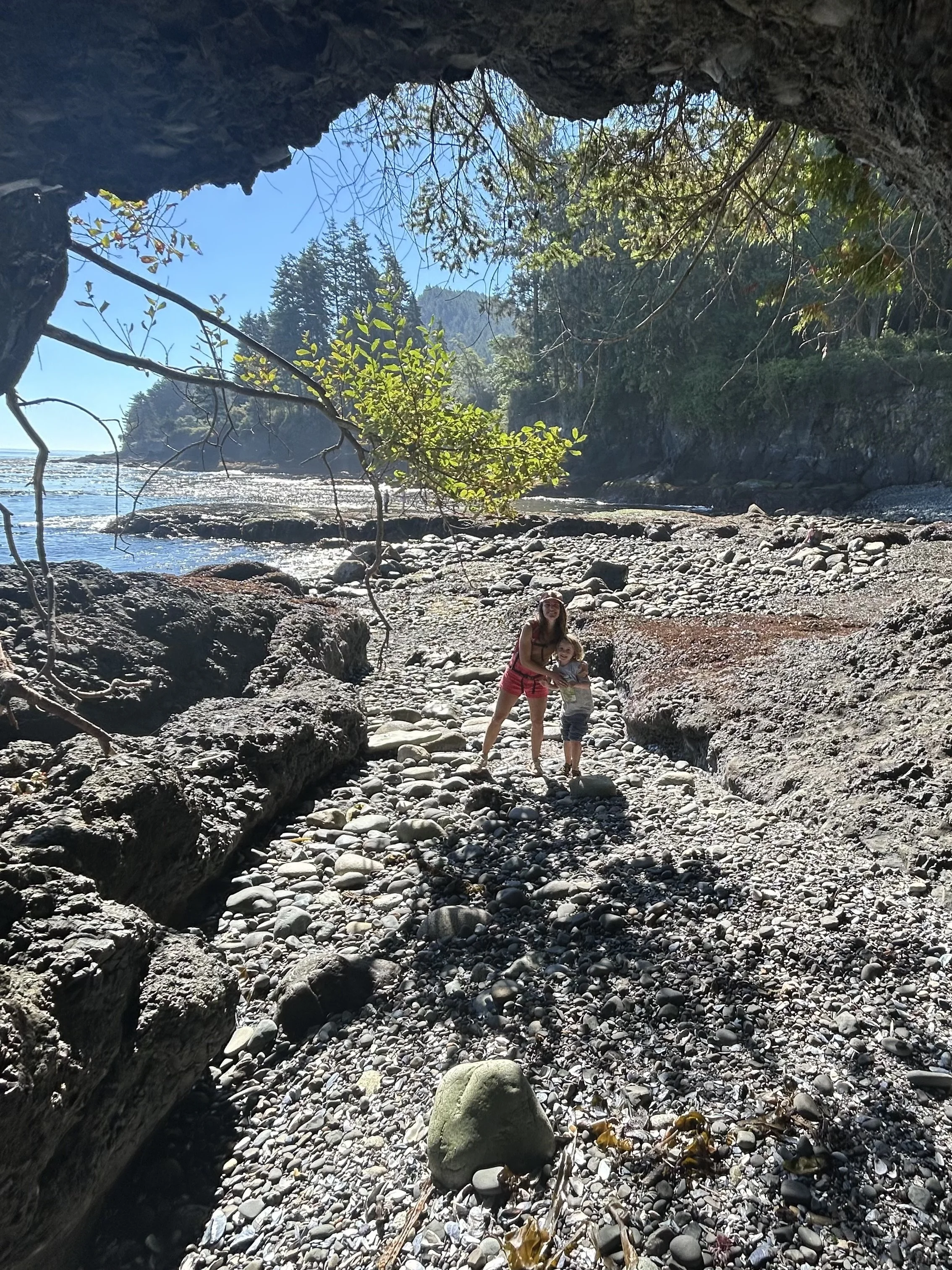 A woman and a young child standing on a rocky beach under a large overhanging rock, with the ocean and forested hills in the background.
