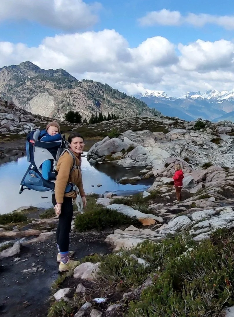 A woman with a child in a backpack carrier standing on a rocky trail by a mountain lake. The woman is smiling and looking at the camera, and a person in a red jacket is looking at the water. There are mountains with snow in the background under a partly cloudy sky.
