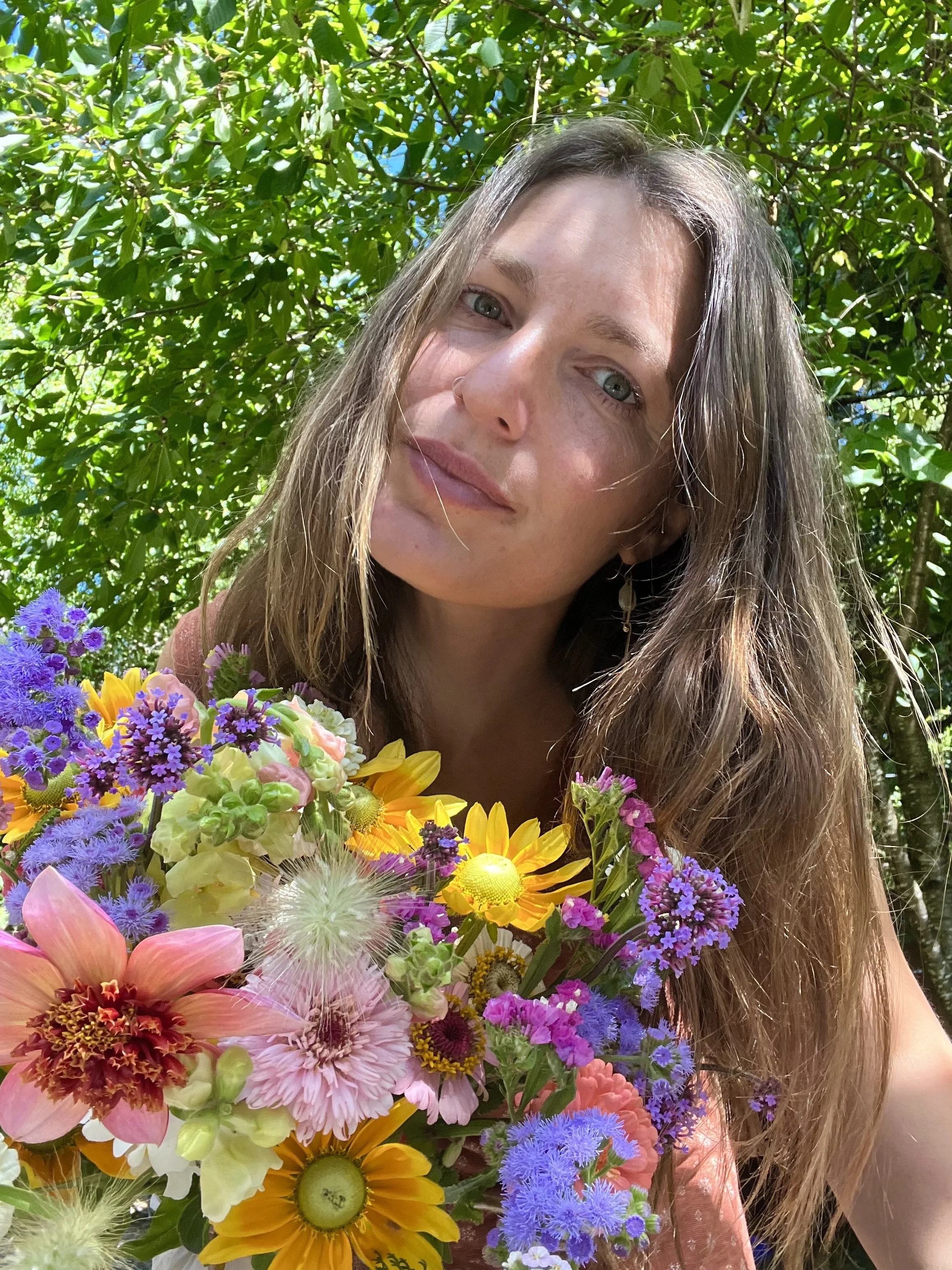 A woman with long brown hair and light eyes holding a colorful bouquet of various flowers outdoors, with green foliage in the background.