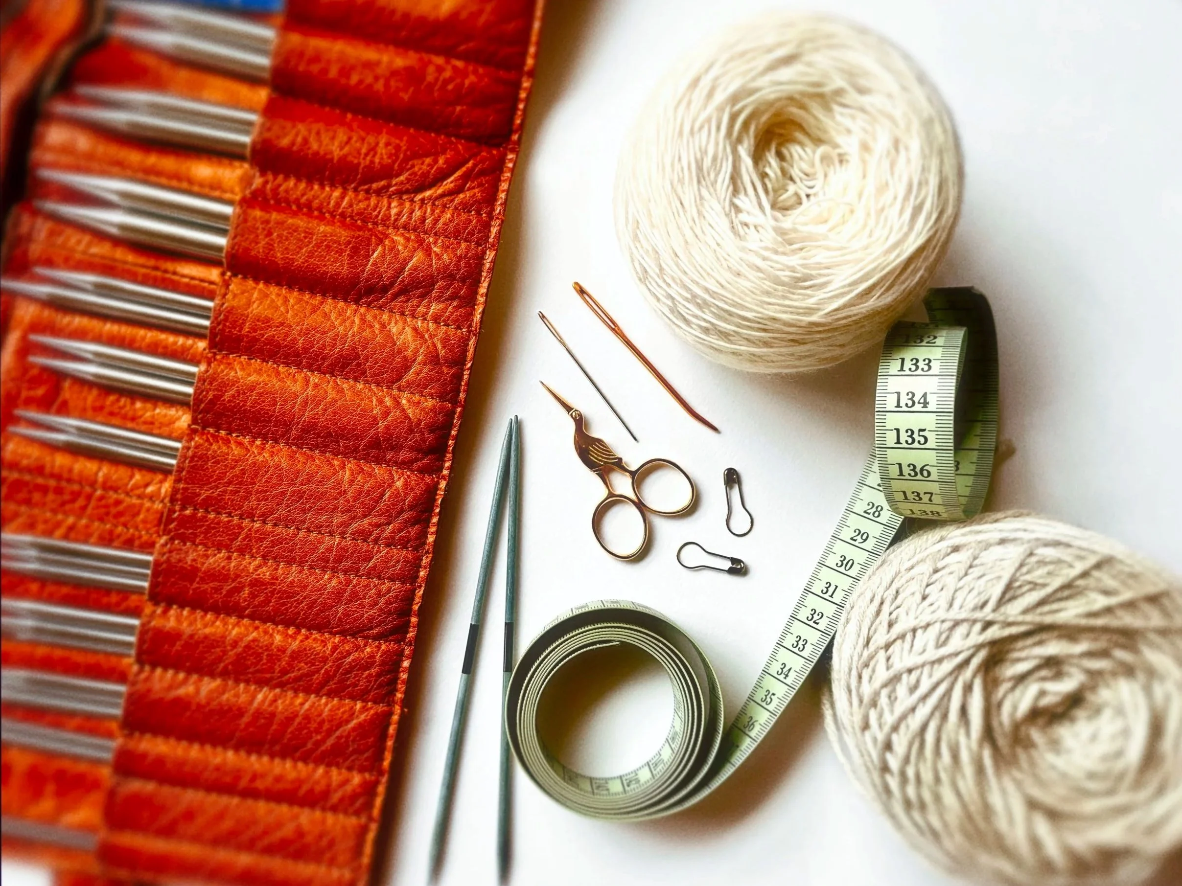 Knitting supplies including orange thread holder with knitting needles, two balls of white yarn, scissors, stitch markers, a measuring tape, and darning needles on a white surface.