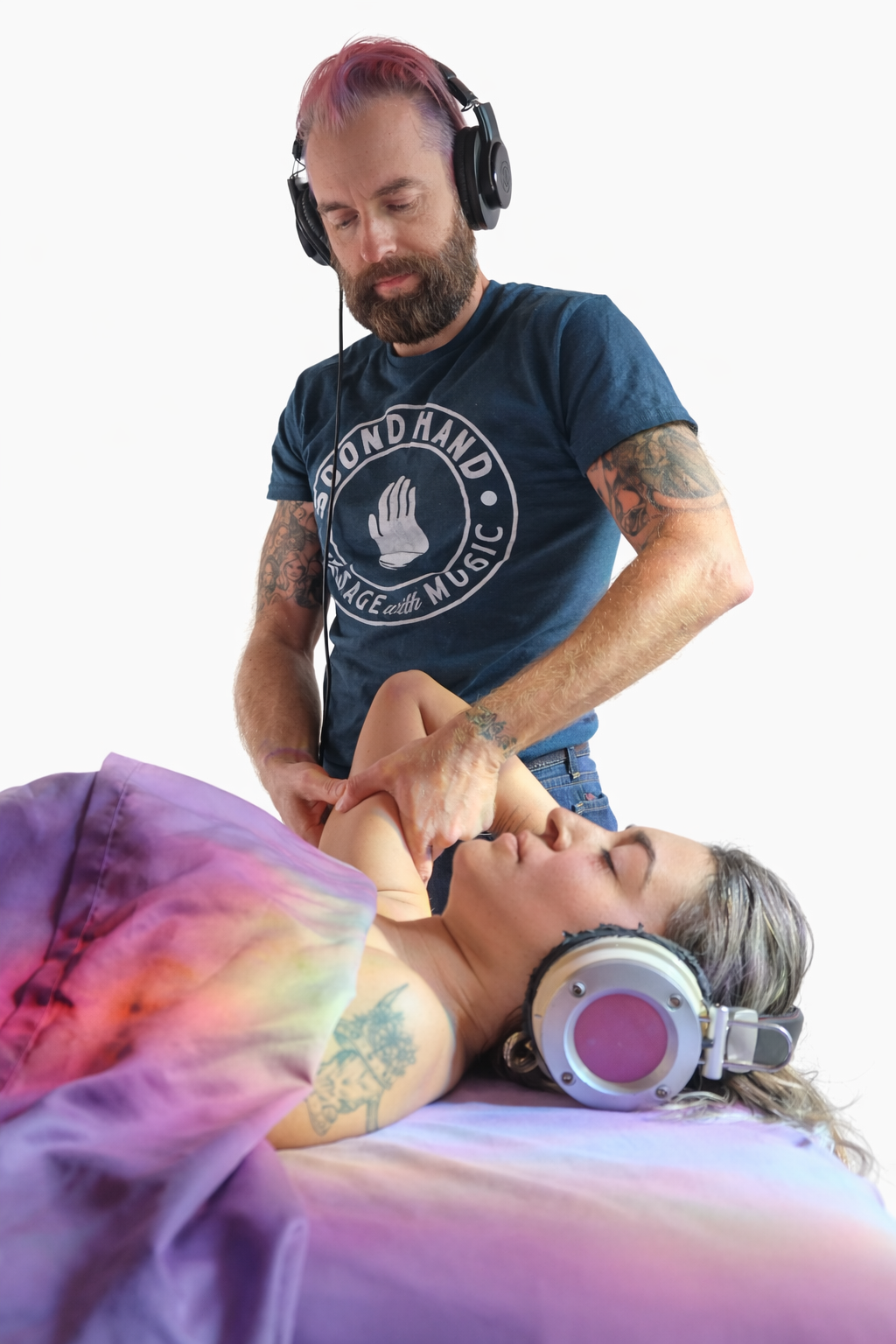 A man with pink hair, a beard, and tattoos is wearing headphones and performing a massage on a woman lying on a massage table, who has gray hair, tattoos, and is wearing headphones.