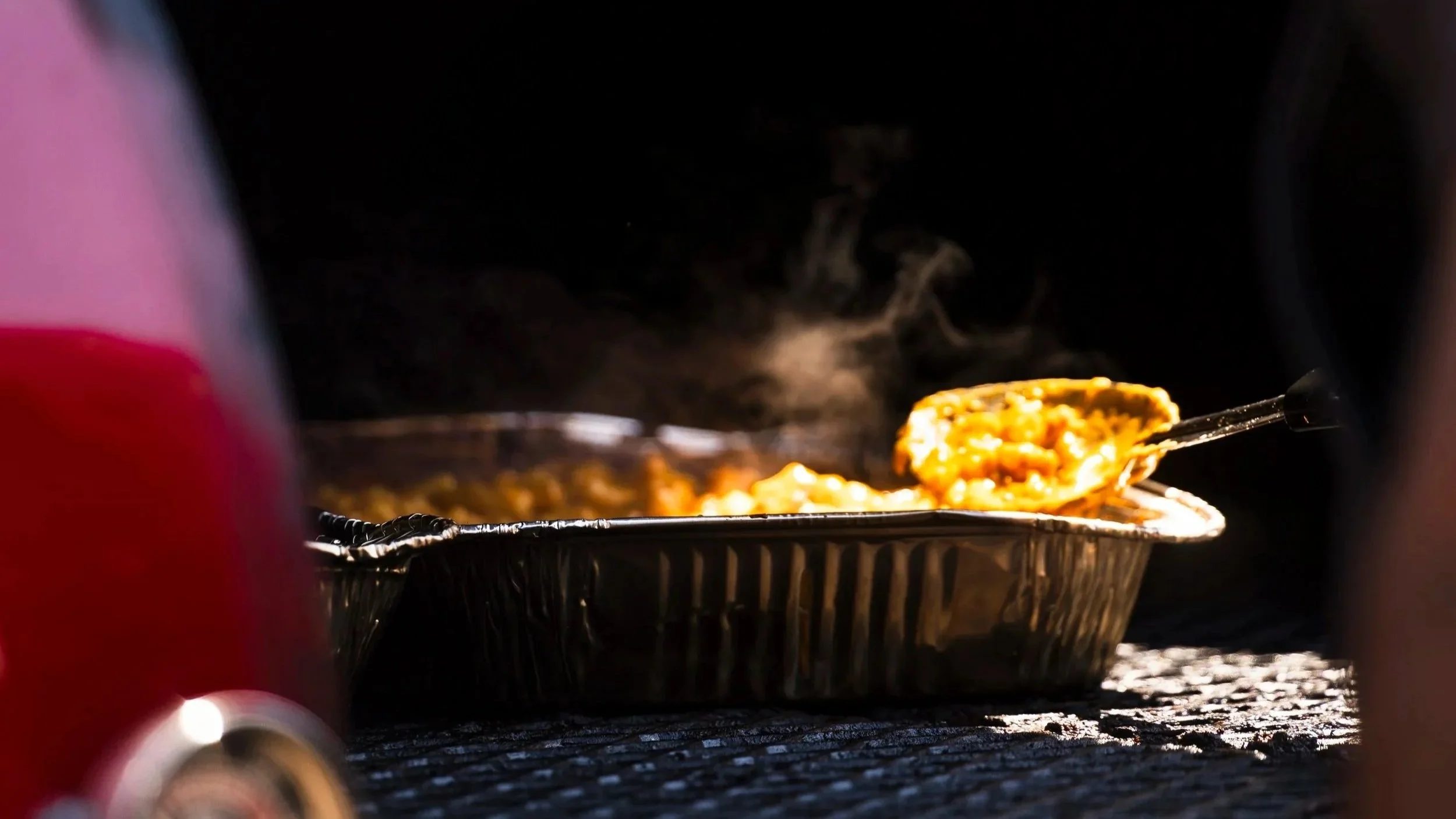 Close-up of food in a takeout container with steam rising, viewed from below, with a person holding tongs.