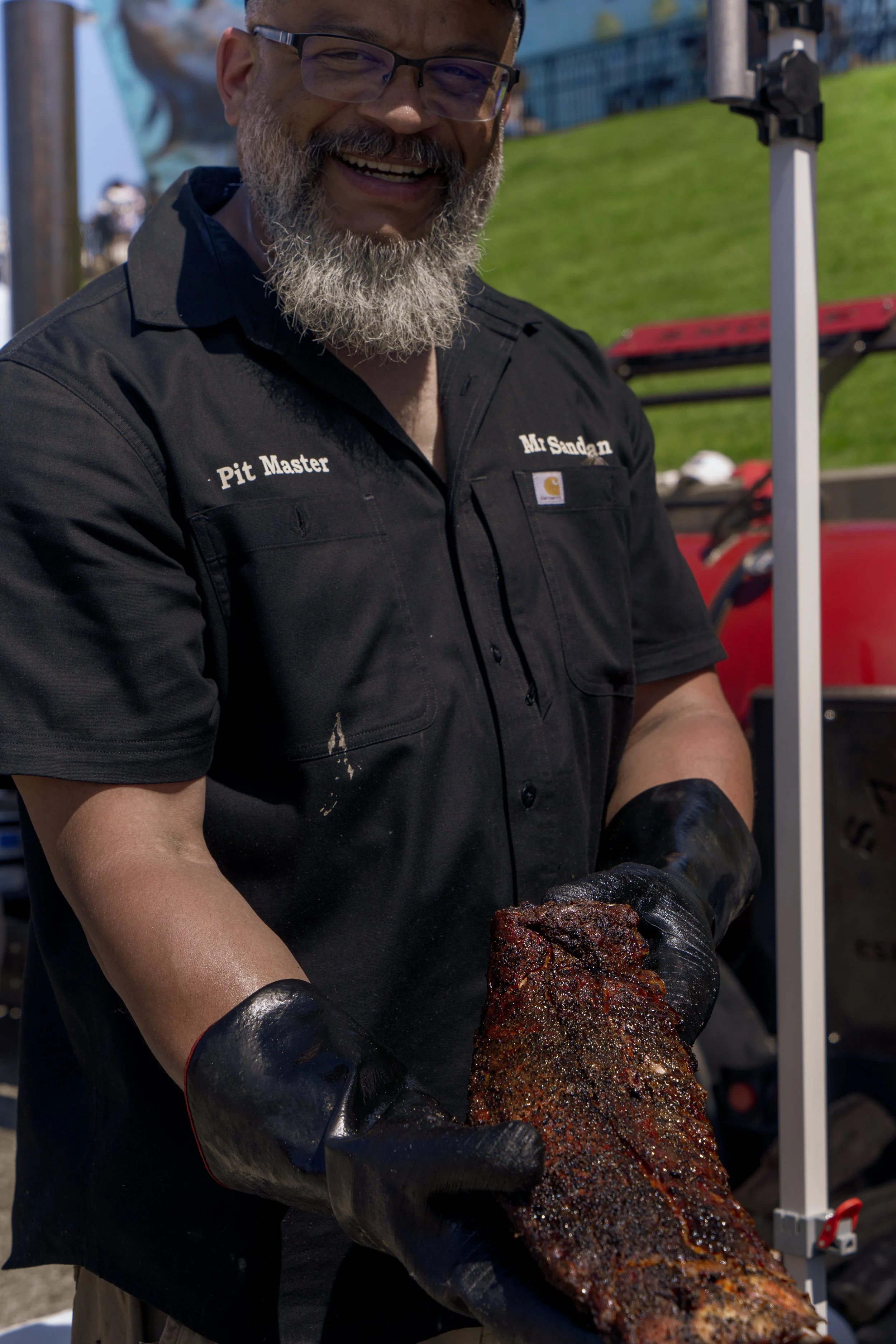 Smiling man with glasses and a gray beard wearing a black shirt with 'Pit Master' and 'Mr. Sandman' embroidered, holding a large piece of grilled meat outdoors.