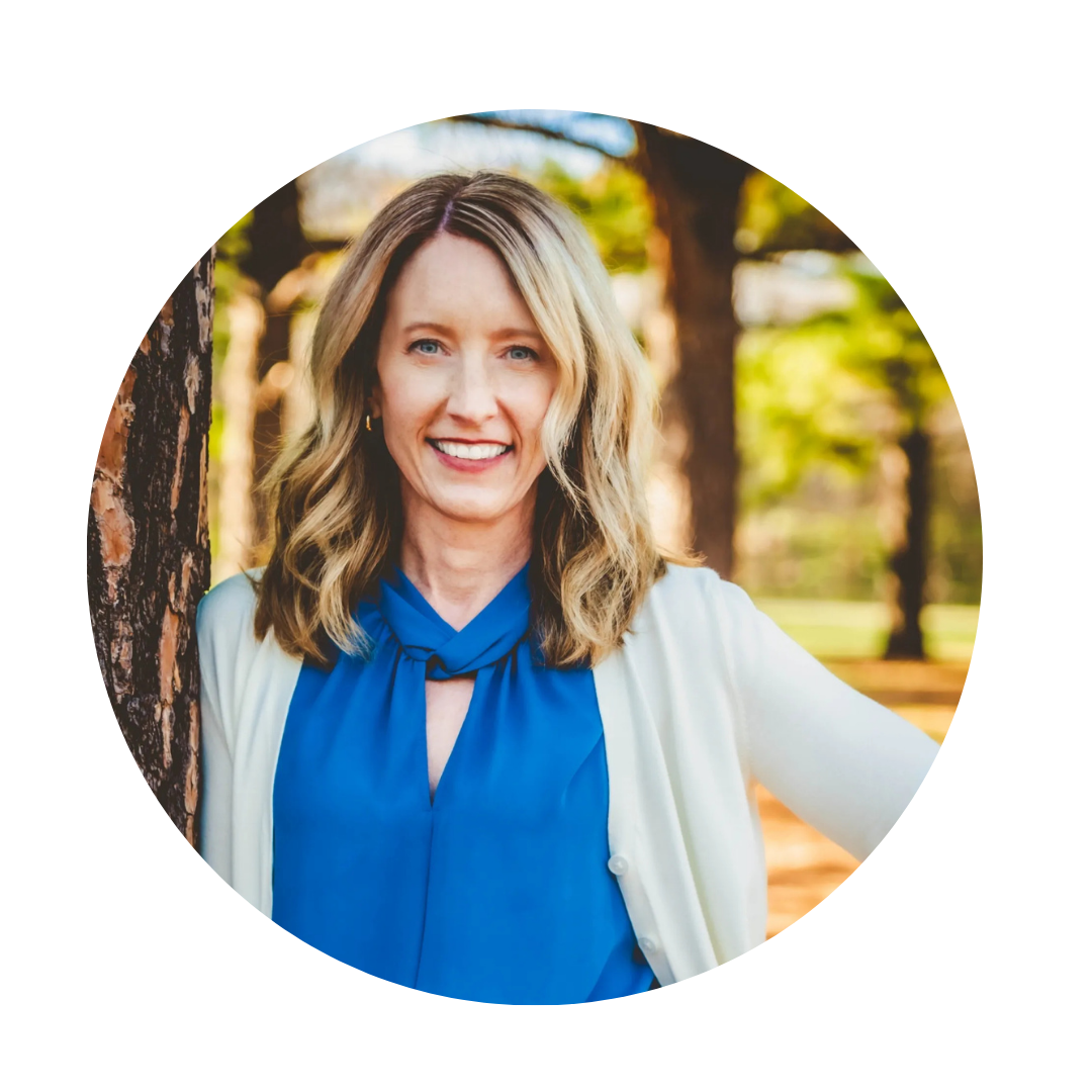 Smiling woman with blonde hair wearing a blue blouse and cream cardigan outdoors in a park with trees in the background.