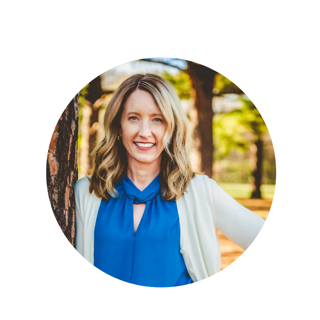 A woman with blonde hair, blue eyes, and a bright smile, standing outdoors among trees, wearing a blue blouse and white cardigan.