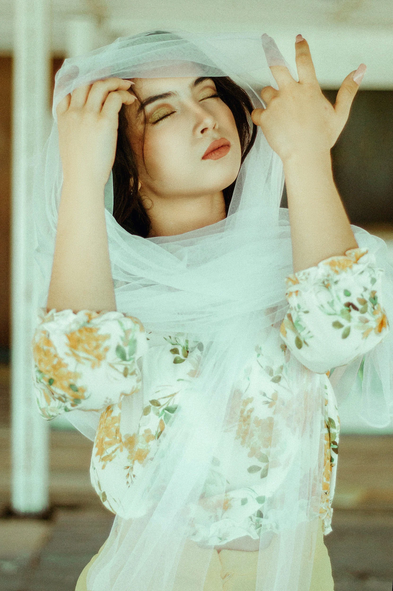 A woman with dark hair and makeup, dressed in a floral blouse with puffed sleeves, is holding sheer white fabric over her head and face, with closed eyes and a serene expression.