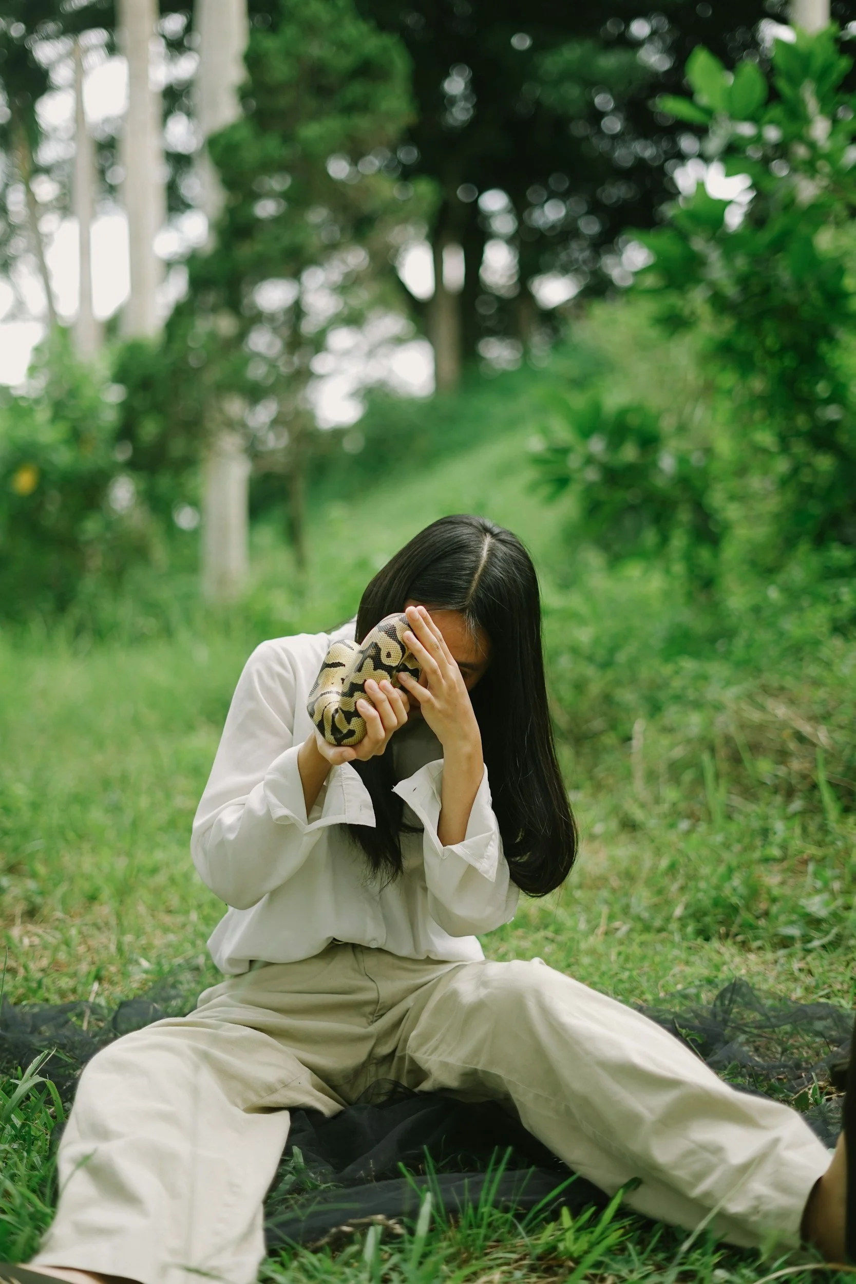 A woman sitting on the grass in a forest, holding a snake close to her face with one hand and covering her eyes with the other.
