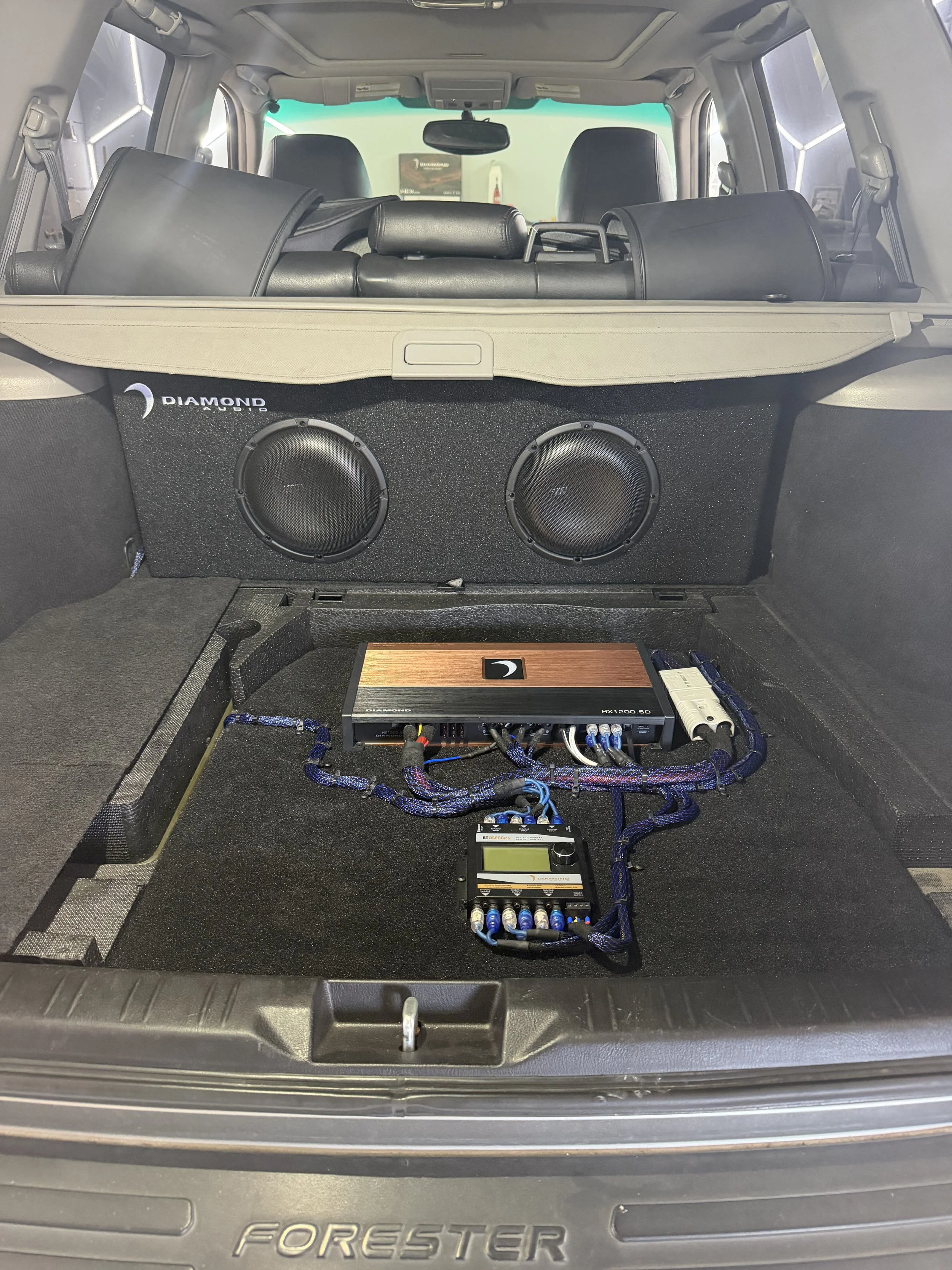 The back of a Ford Foresster vehicle with a large black subwoofer and audio equipment installed in the trunk. The trunk is empty except for the audio system and wiring.