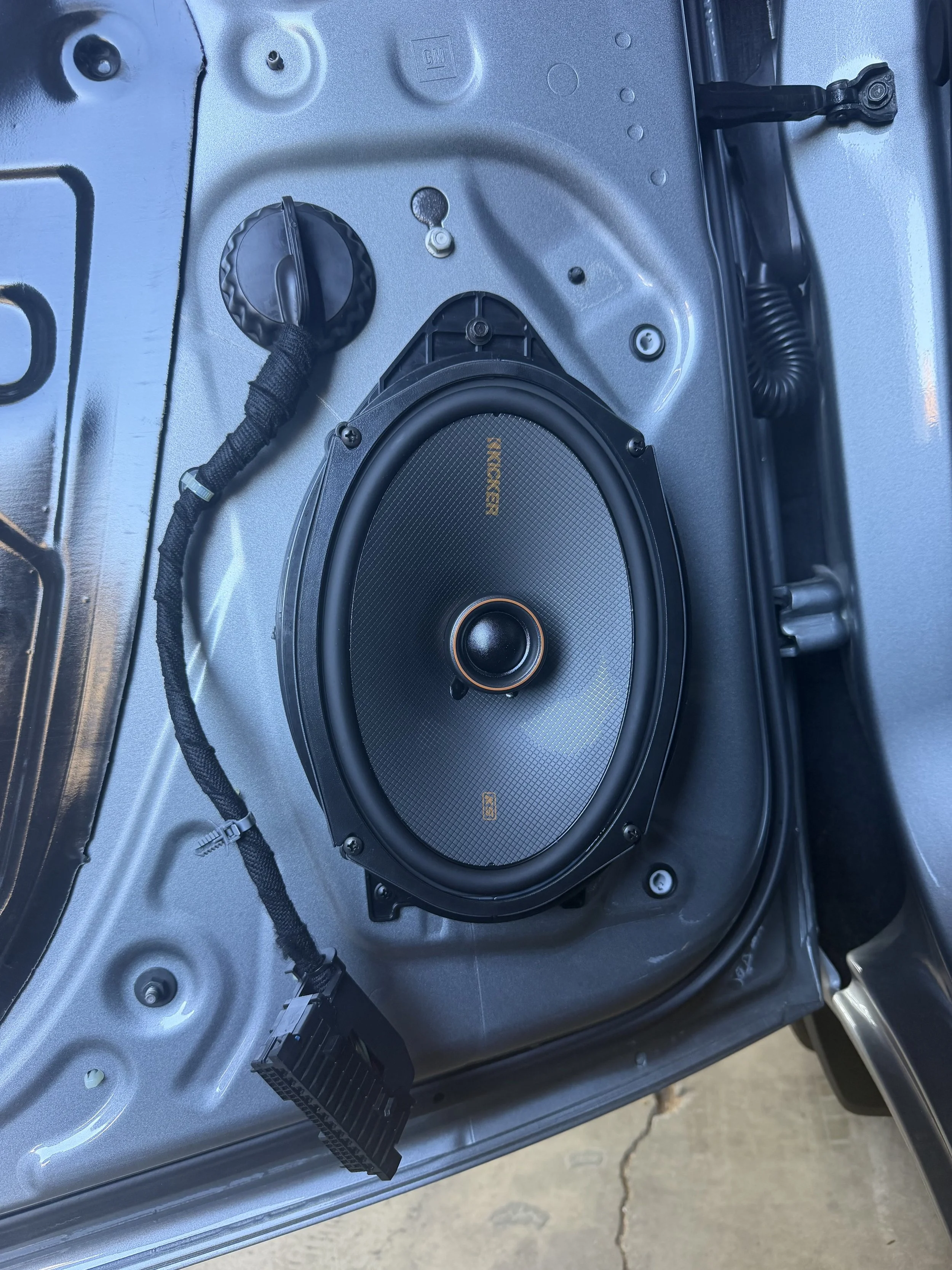 Close-up of a vehicle's interior showing a mounted speaker, electrical wiring, and a metal panel with screws, with a cracked concrete ground outside.