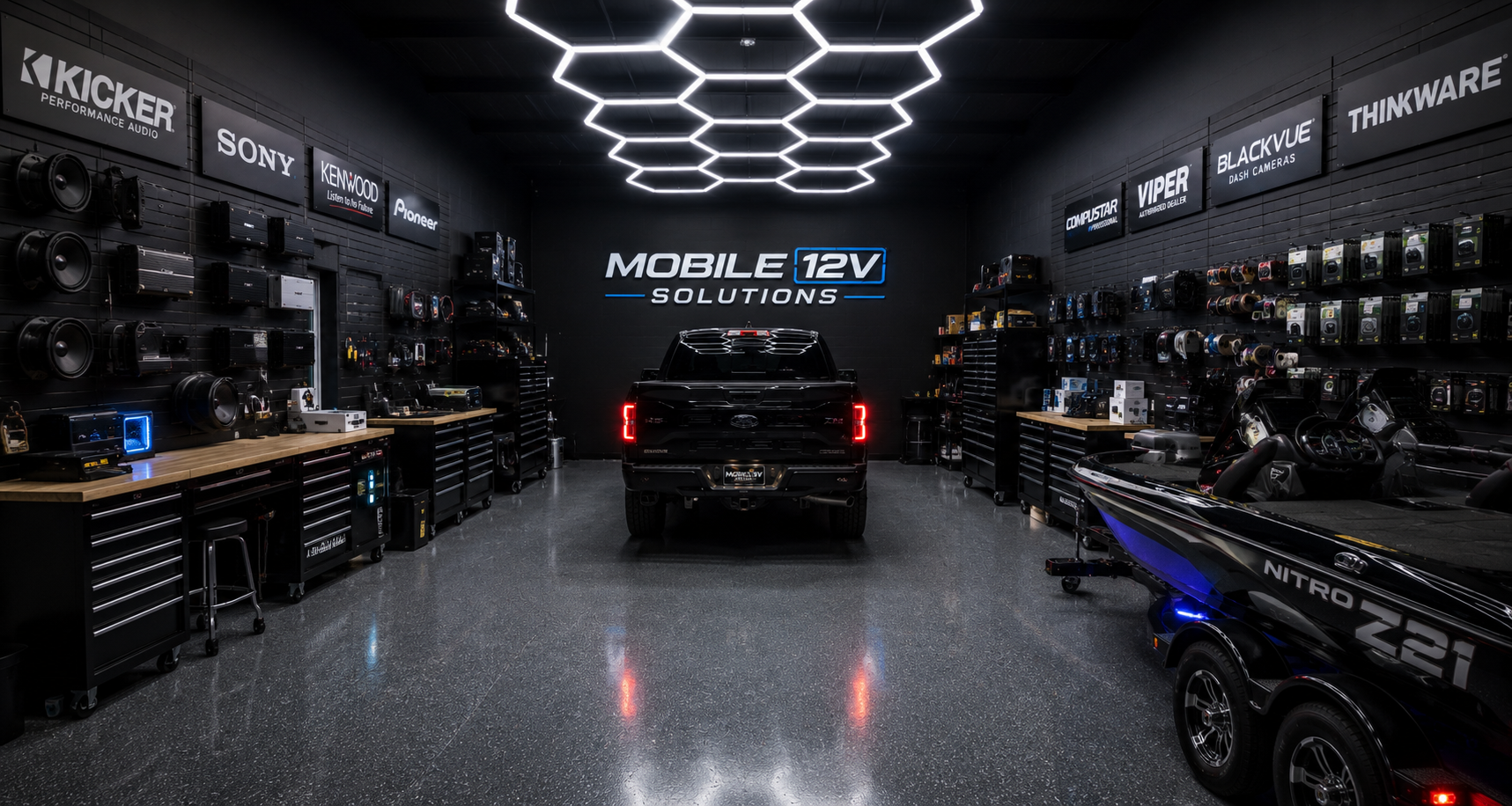 Interior of an automotive electronics and accessories store with a black pickup truck in the center, surrounded by various brands of audio equipment, dash cameras, and other vehicle tech, with a neon sign reading 'MOBILE 12V SOLUTIONS' on the back wall.