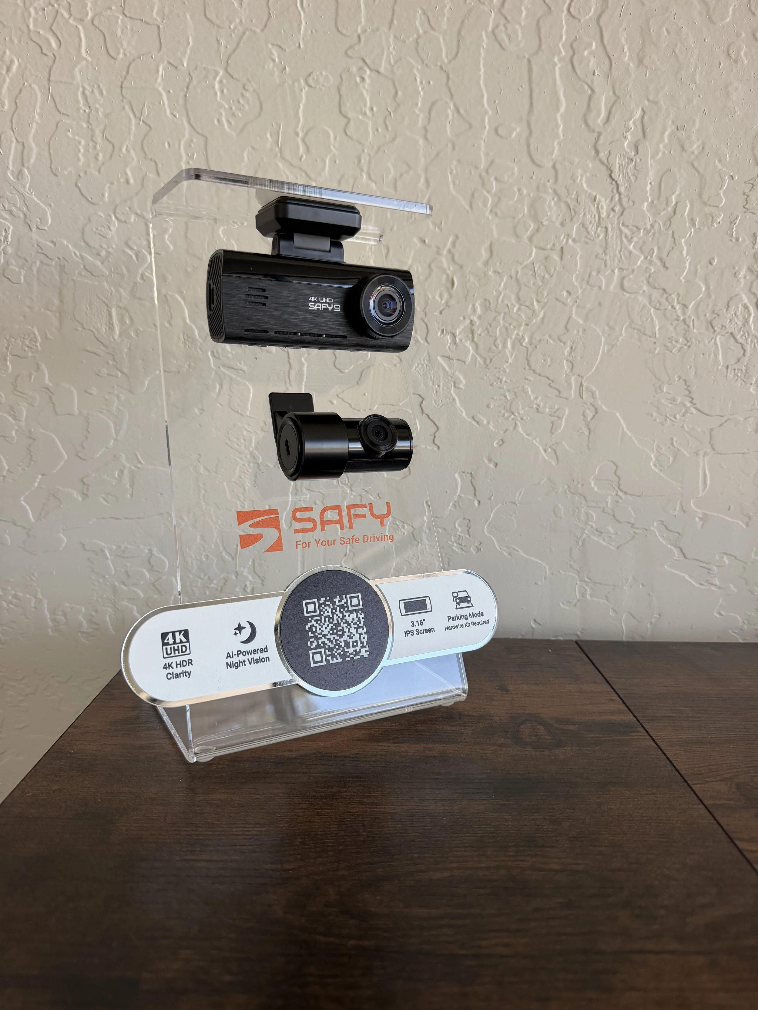 Display stand showcasing two dash cameras on a wooden surface with a beige textured wall in the background, branded with 'SAFEY' and features specifications including 4K UHD, AI-powered night vision, 3.16-inch IPS screen, and parking mode.