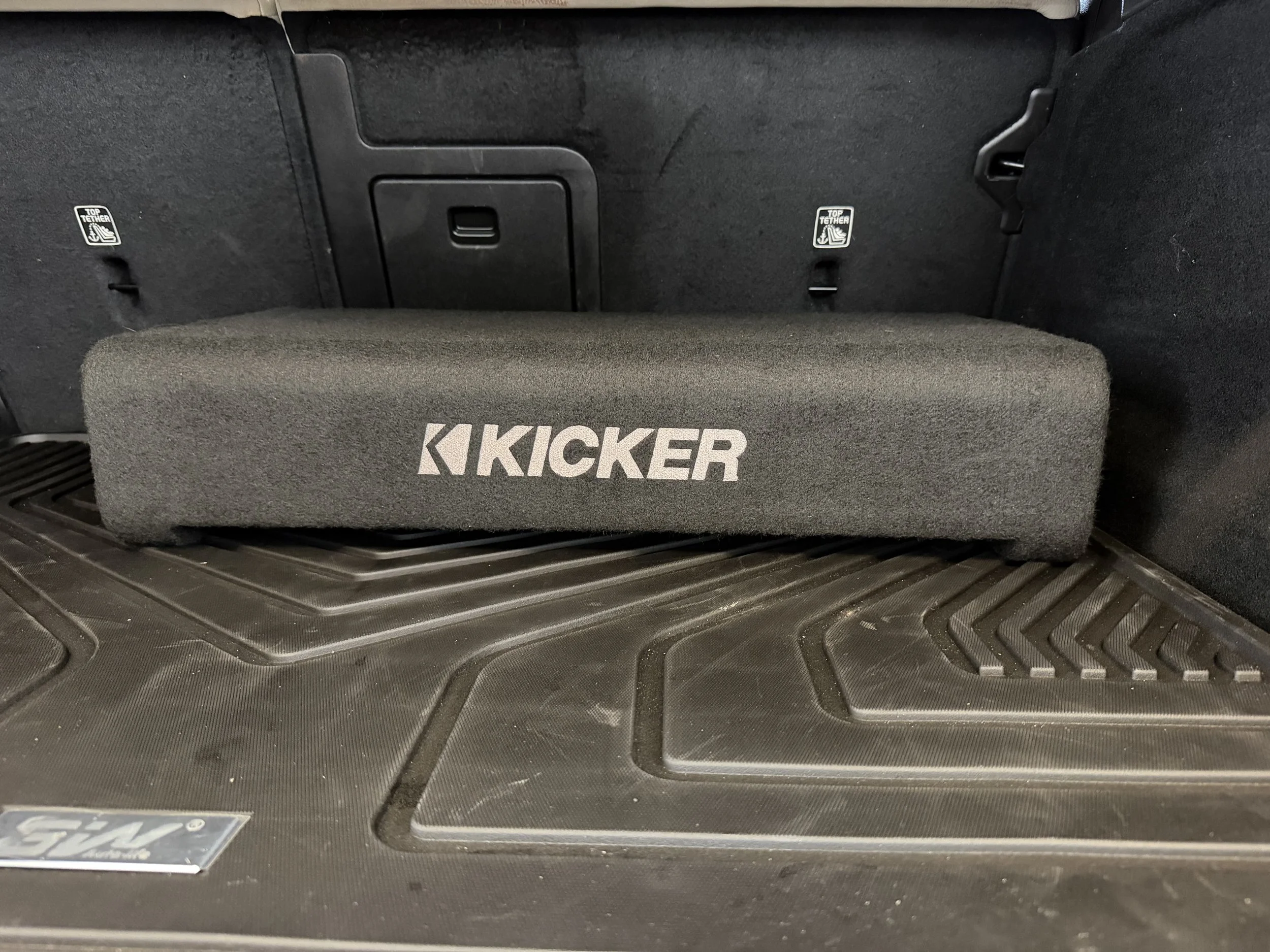 The image shows the interior trunk space of a vehicle with a carpeted cargo cover featuring the Kicker brand logo, and plastic panels with labels indicating where to attach top tether anchors.