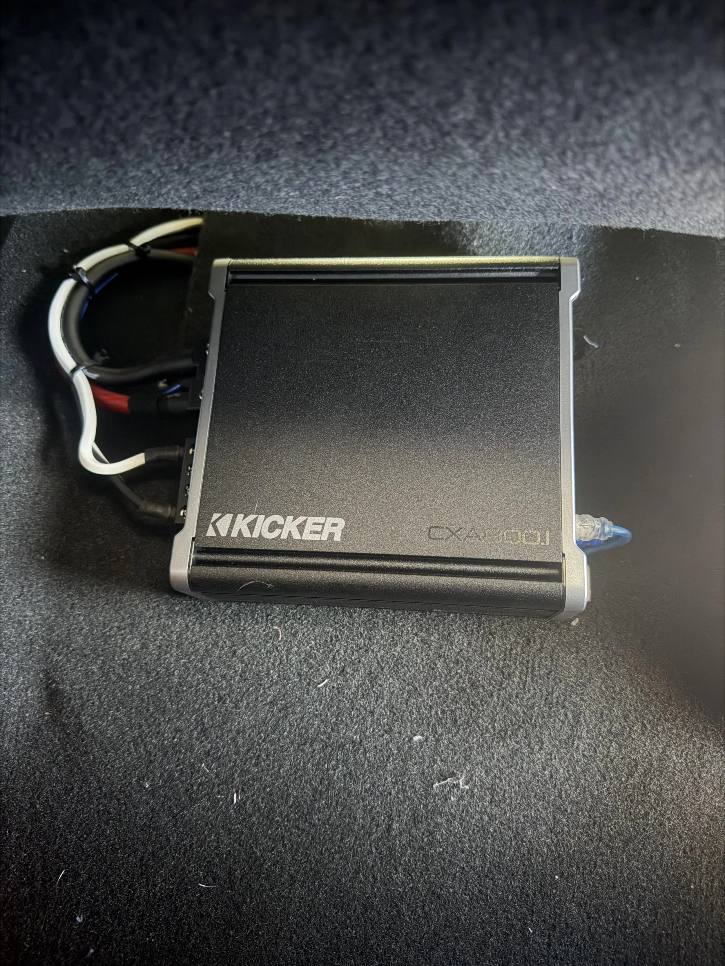 A black and silver Kicker CXA AUTO 1 amplifier on a black textured floor with cables connected to it.