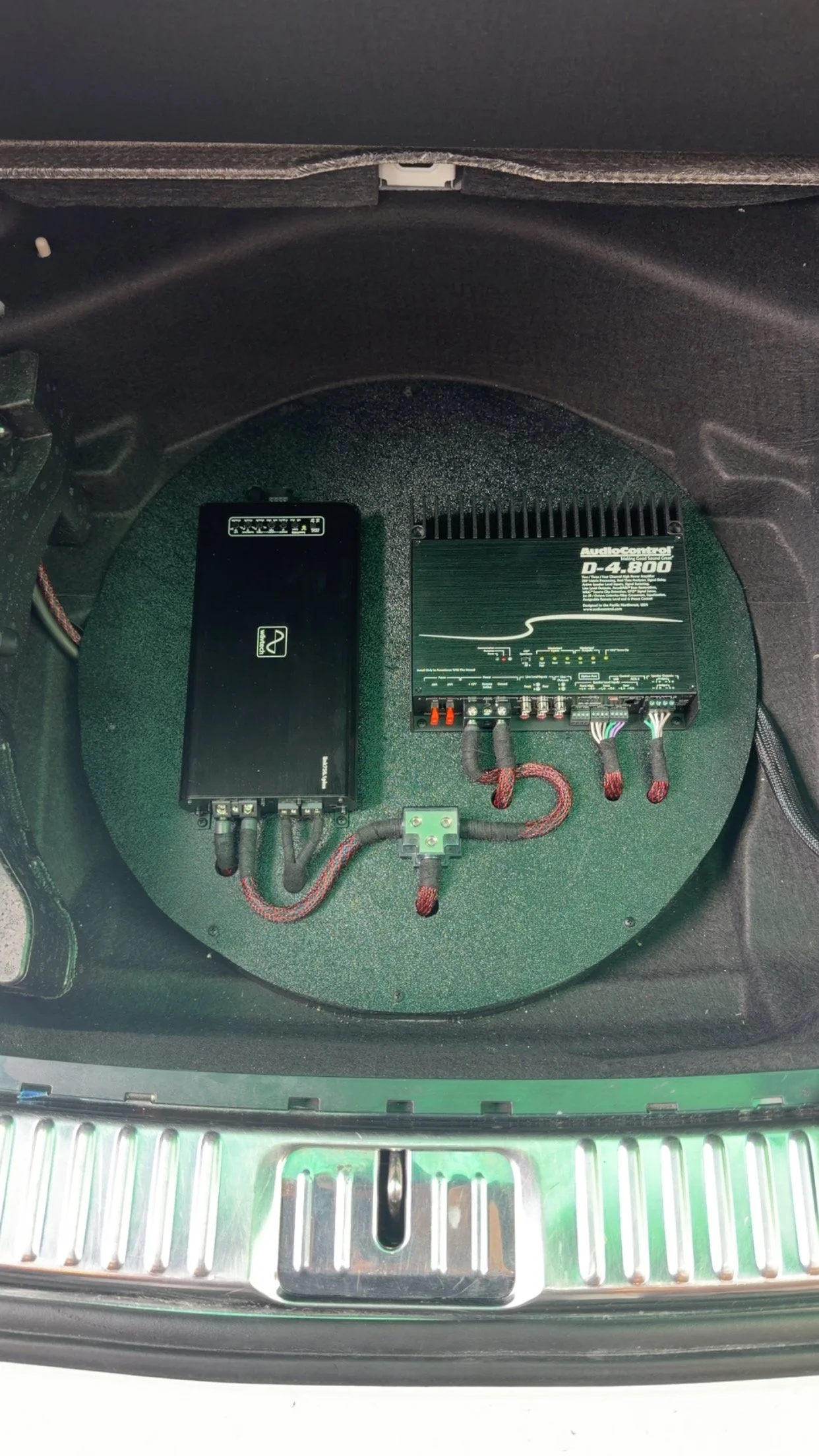 Car trunk with audio amplifier, signal processor, and wiring installed on a green speaker platform.
