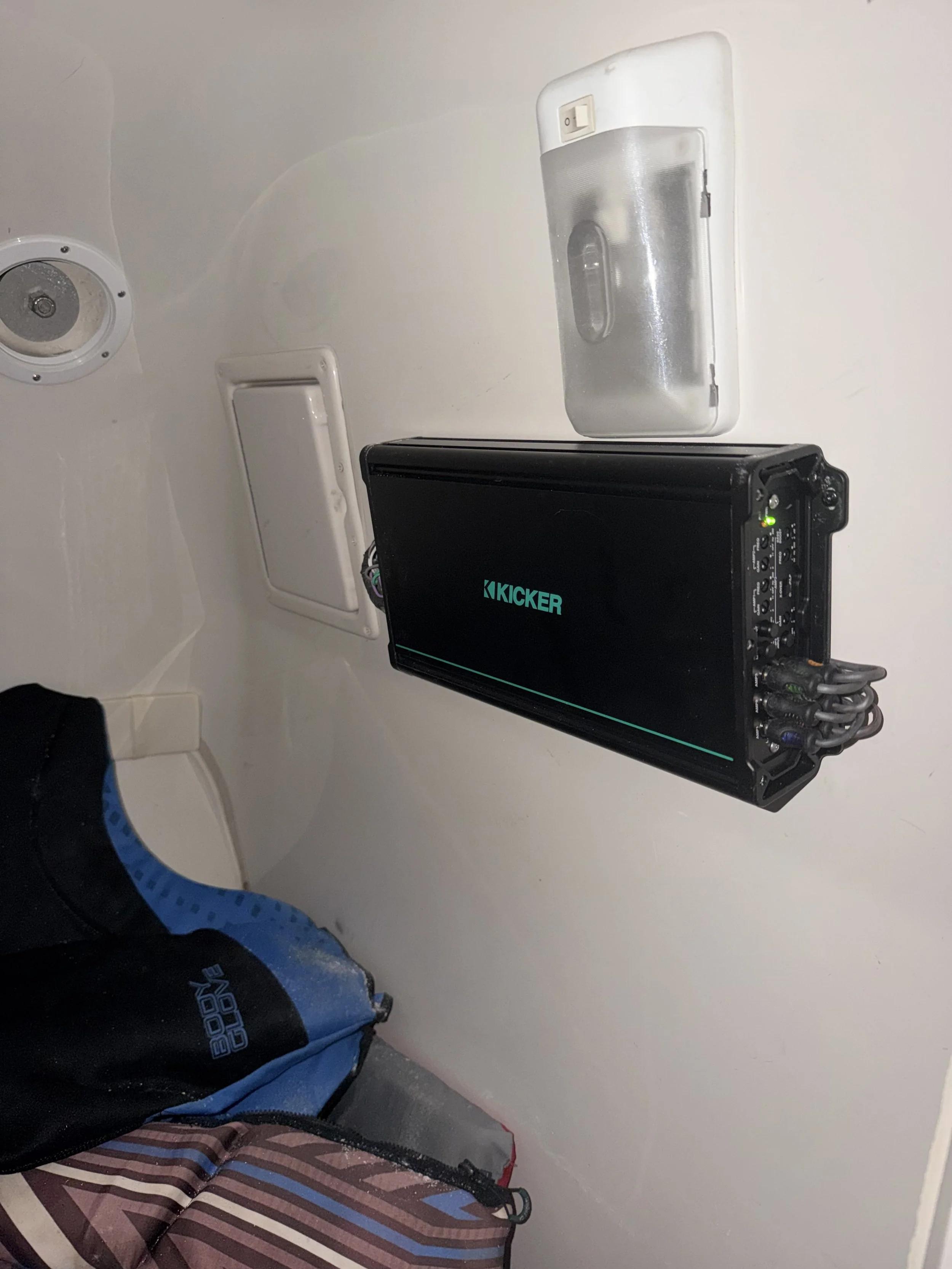 A wall with a ceiling speaker, a small white vent cover, a control switch, a silver rectangular ice chest, a black and blue backpack, and a black electronic device with the word 'KICKER' on its screen.
