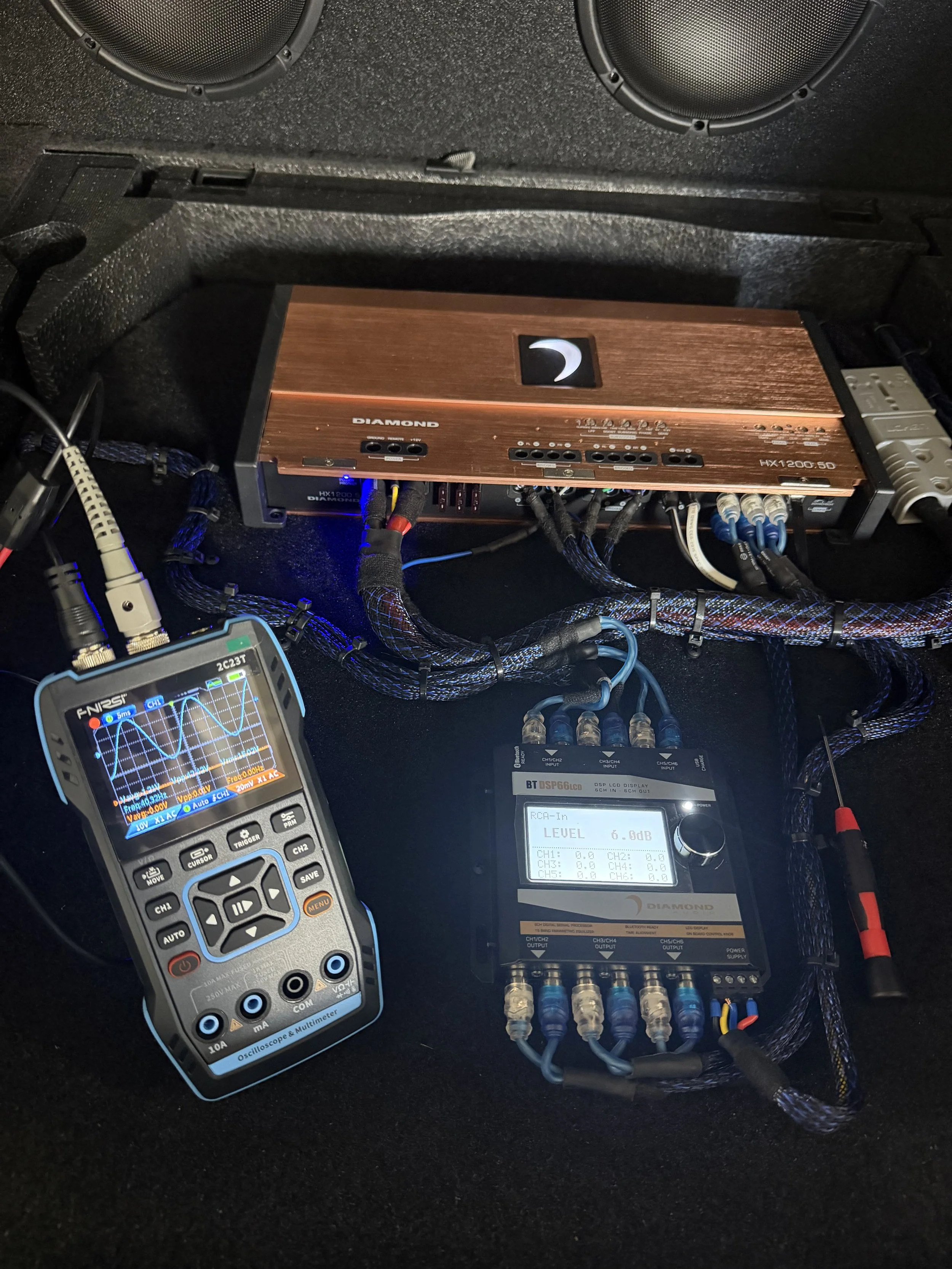 Photo of audio equipment setup with various devices, including a multimeter, an amplifier, and labeled cables connected in a car or sound system environment.