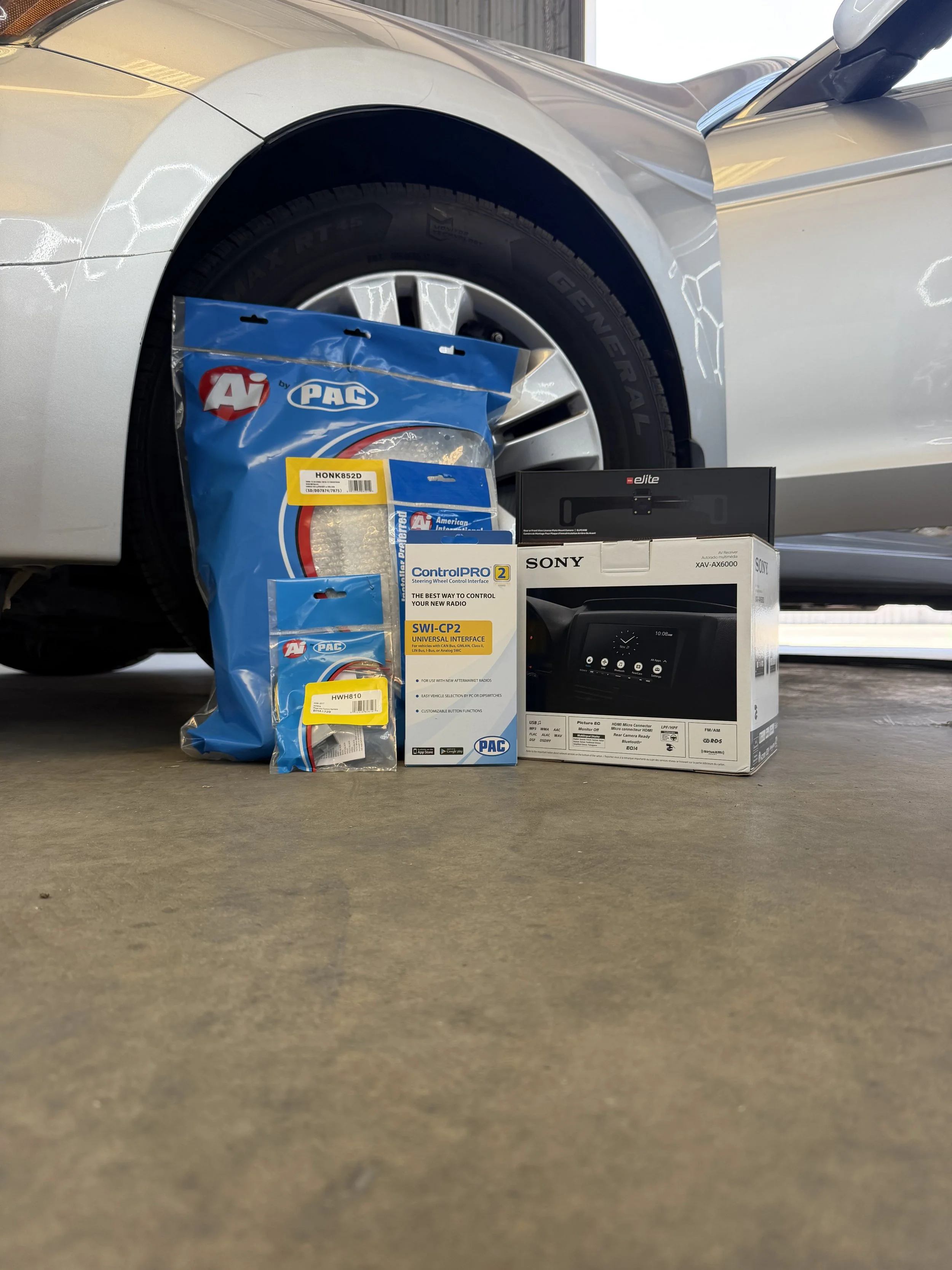 The image shows the front wheel of a silver car parked in a garage or warehouse, along with various electronic and automotive items placed on the concrete floor in front of the wheel.