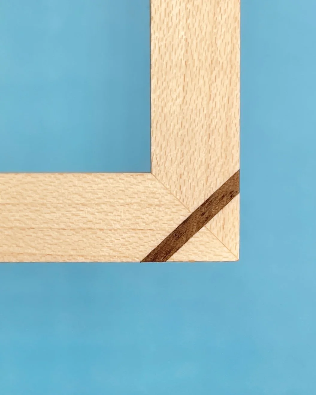 Detail eines Ahornrahmens (Ecke) mit Eckverstärkung aus Nussbaumholz vor blauem Hintergrund.