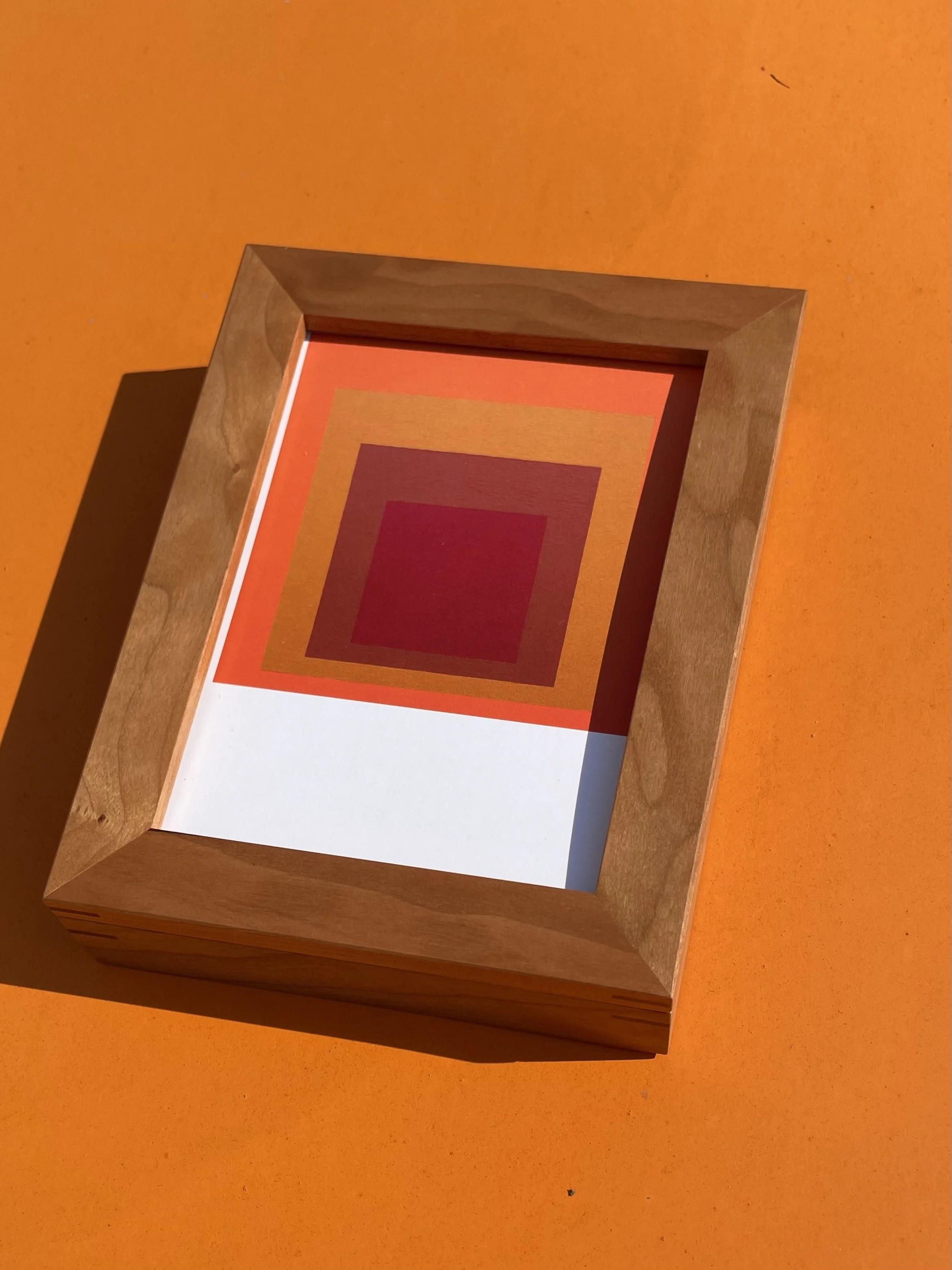 Ein Kirschholzrahmen, darin eine Postkarte mit einem Motiv von Josef Albers, das aus mehreren Rechtecken in verschiedenen Rot- und Orangetönen besteht. Liegt auf einem orangefarbenen Untergrund.
