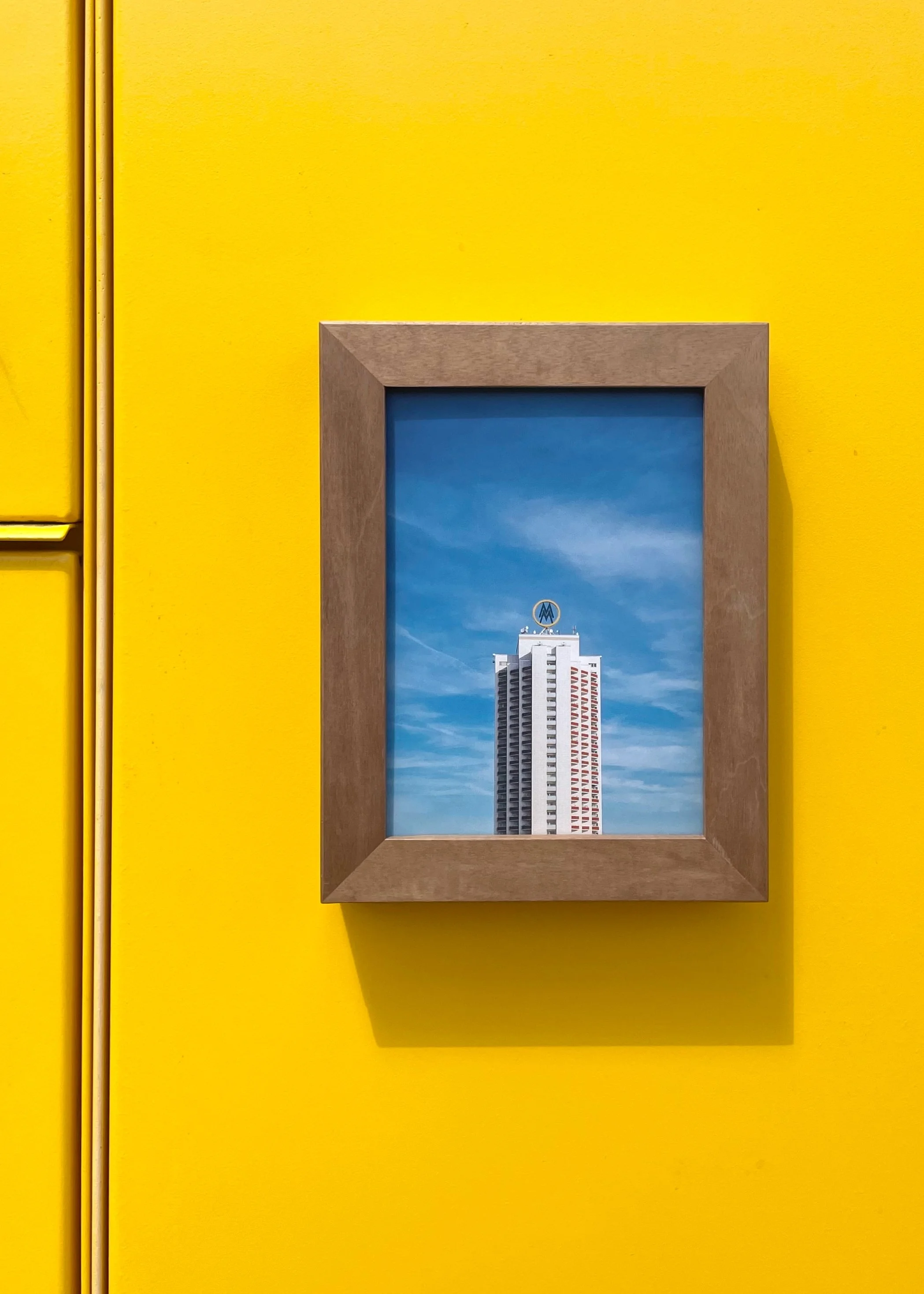 Ein Nussbaum-Rahmen, der eine Postkarte des 'Messe'-Towers in Leipzig zeigt. Vor gelbem Hintergrund, vermutlich eine DHL-Packstation.