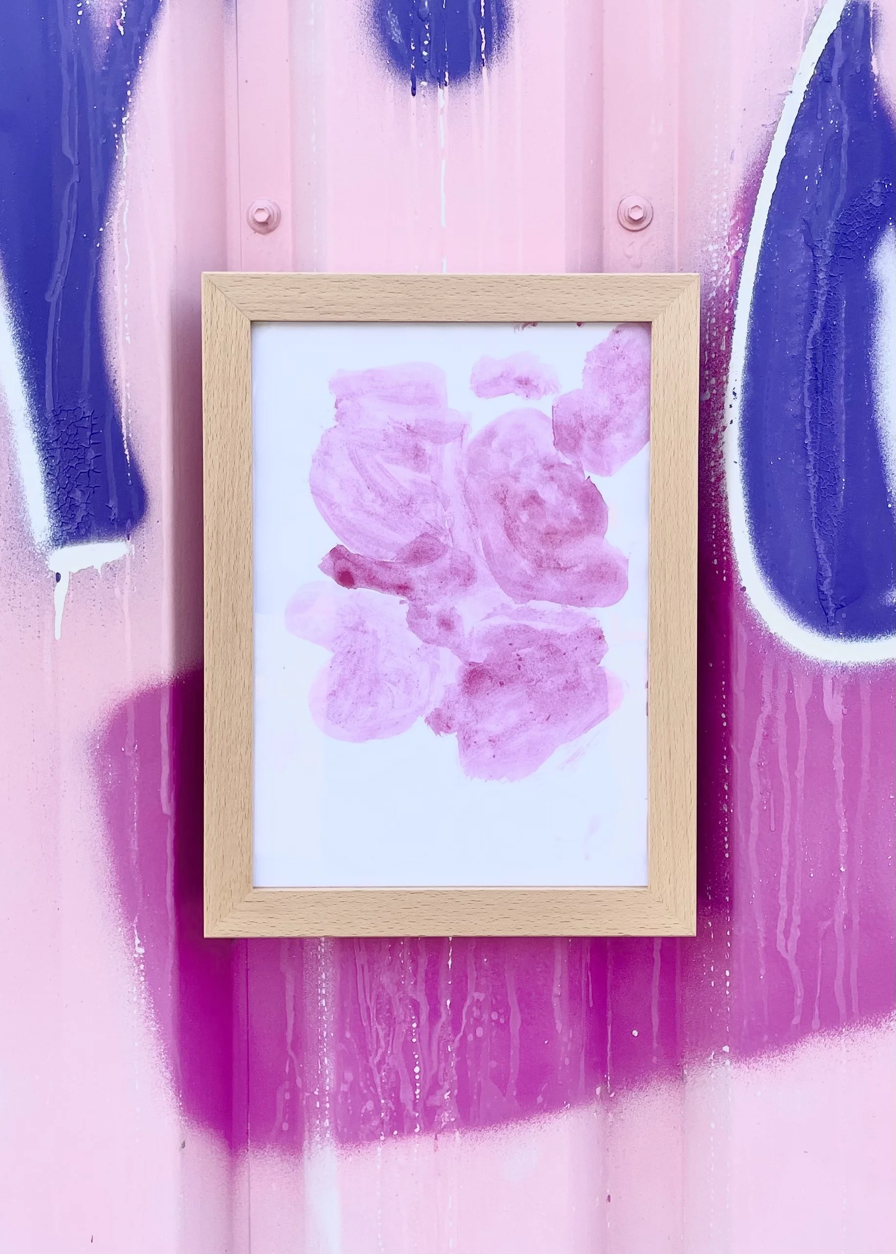 Abstraktes Kinder-Kunstwerk mit pinken Farbverläufen in hellem Buchenholz-Rahmen, vor einer Hauswand mit Graffiti aus blauen, lilanen und rosanen Farbflächen.