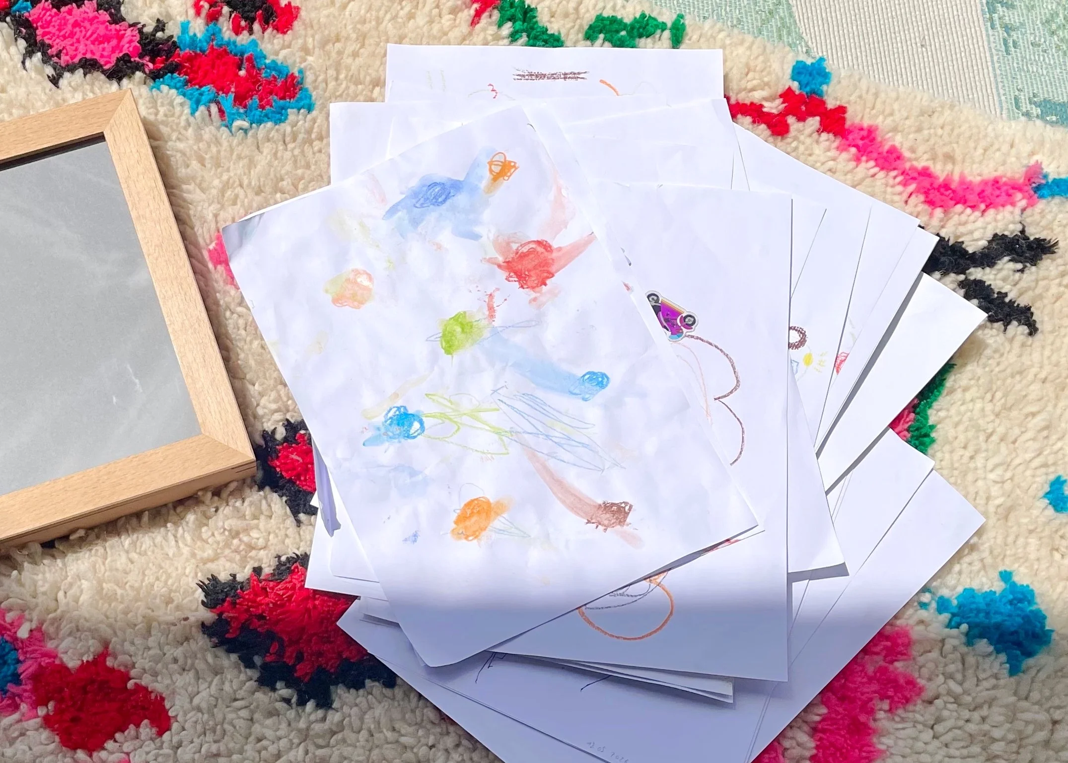 Bunte Kinderzeichnungen, die auf einem Teppich liegen, teilweise mit Stickern beklebt, neben einem leeren Bilderrahmen.