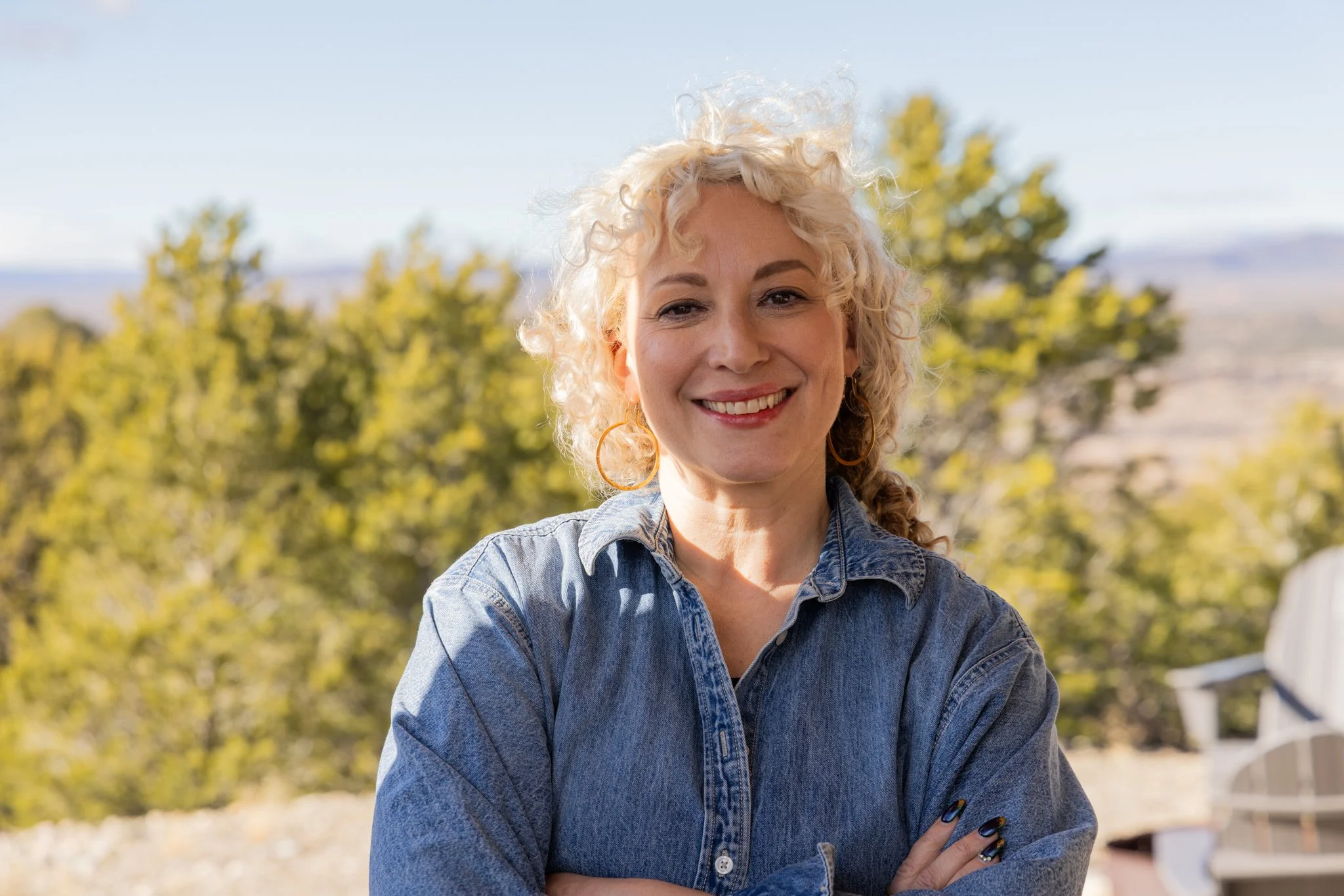 A woman with curly blonde hair smiling outdoors, wearing a denim shirt, with trees and a clear sky in the background.