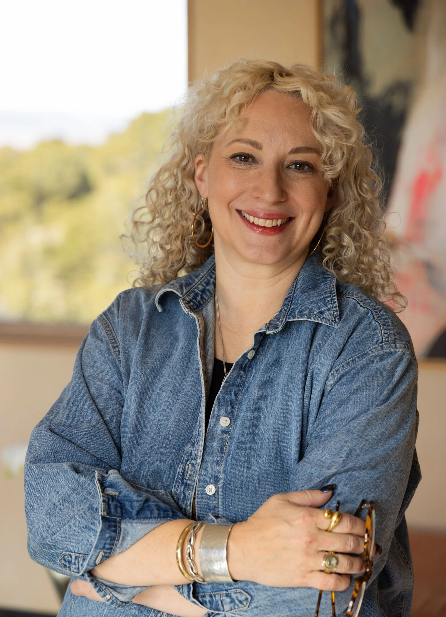 A woman with blonde curly hair, wearing a denim shirt and jewelry, smiling with her arms crossed, in a well-lit indoor space with a window and artwork in the background.