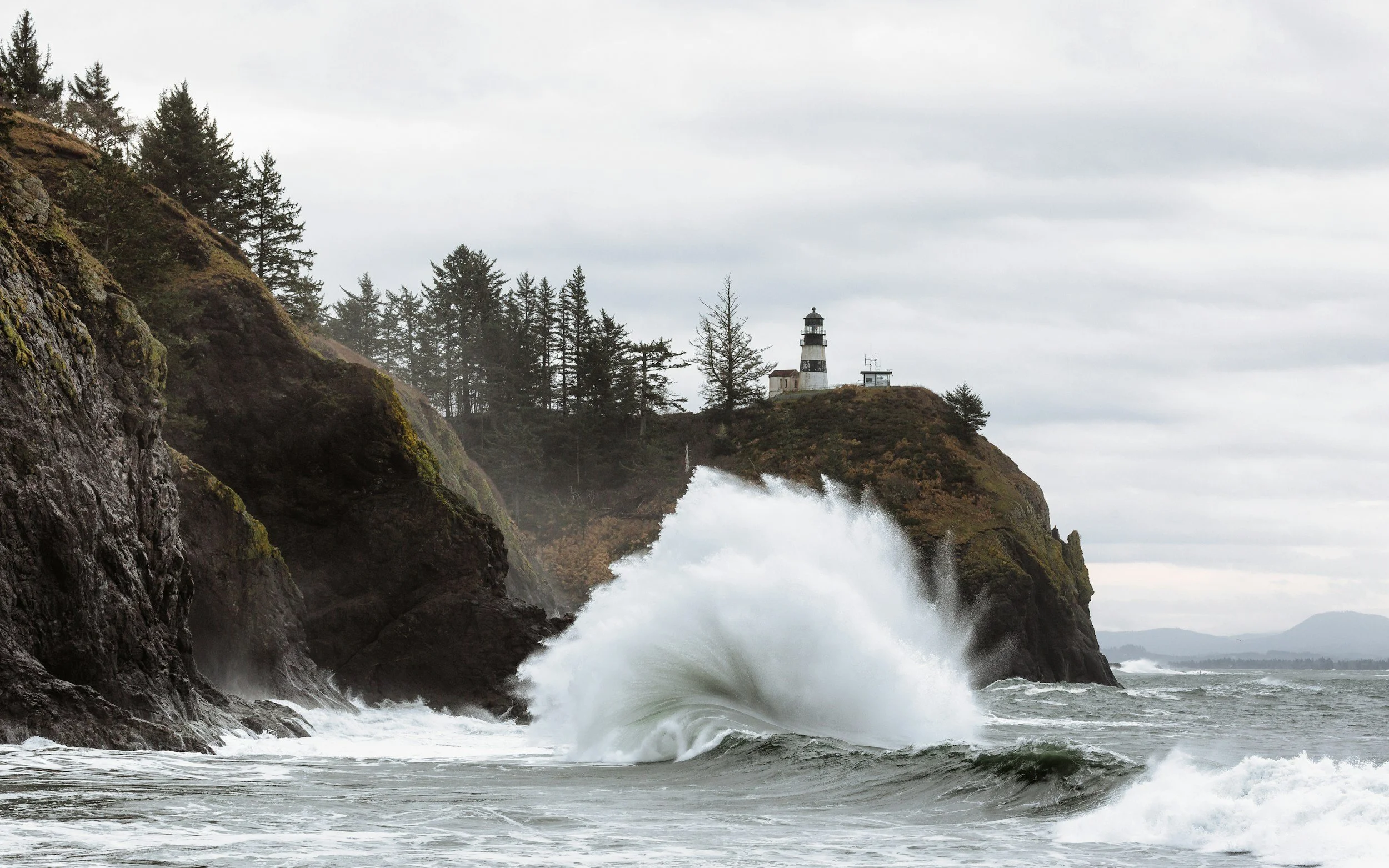 Waves crashing against rocky coastline with a lighthouse on a hill in the distance under cloudy sky.
