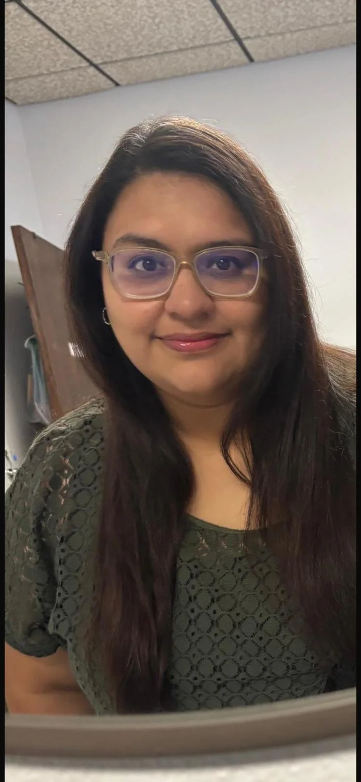 A woman with long dark hair, glasses, and a green lace top smiling at the camera in an indoor setting.