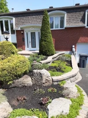 Une maison a Saint-Bruno avec des plates-bandes remplies de fleurs, de plantes, de paillis, de buissons, avec un cèdre et des pavés.