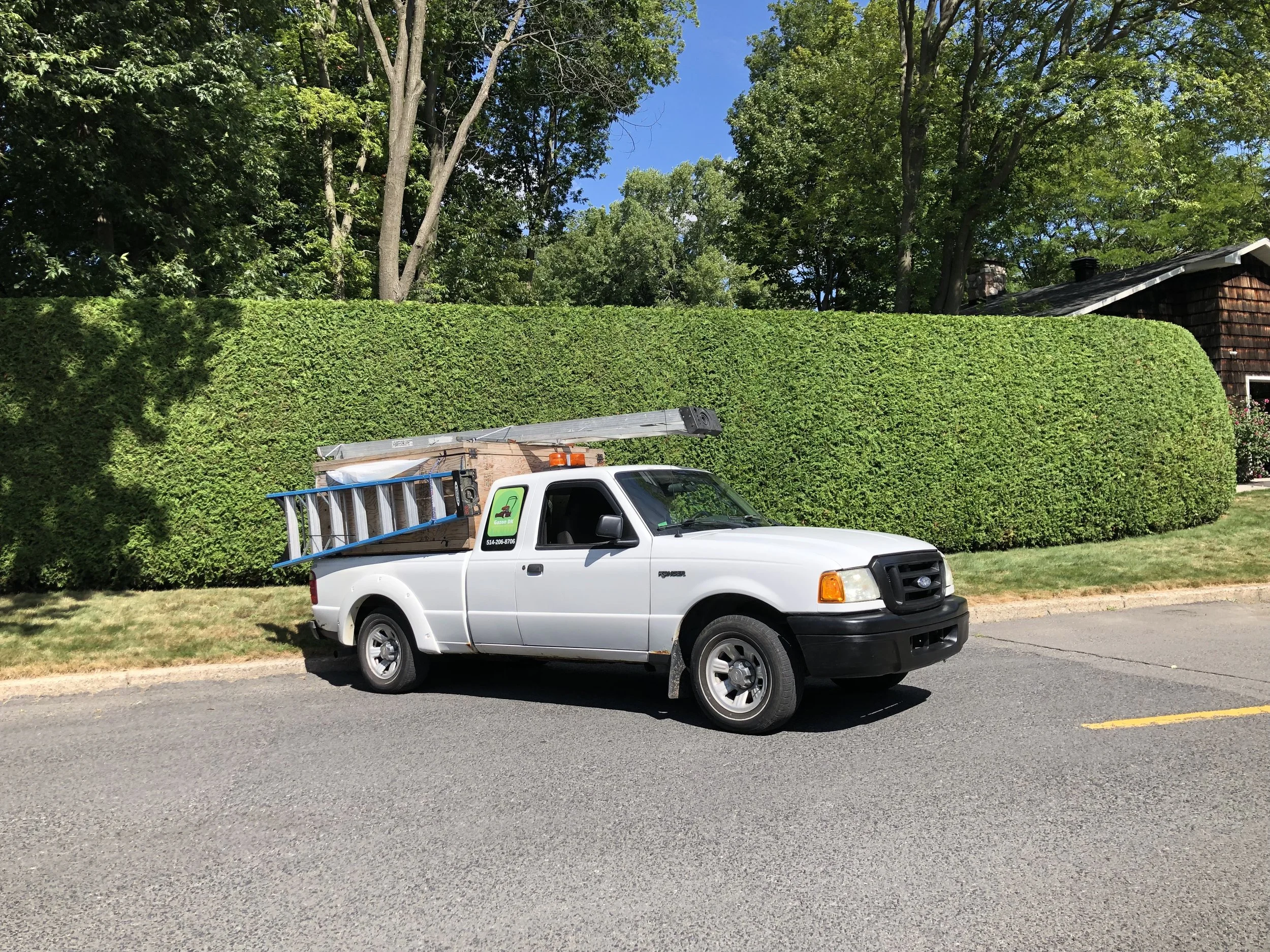 Un camion de paysagement avec de l'équipement devant une haie de cèdres bien taillée à Saint-Basile-le-Grand