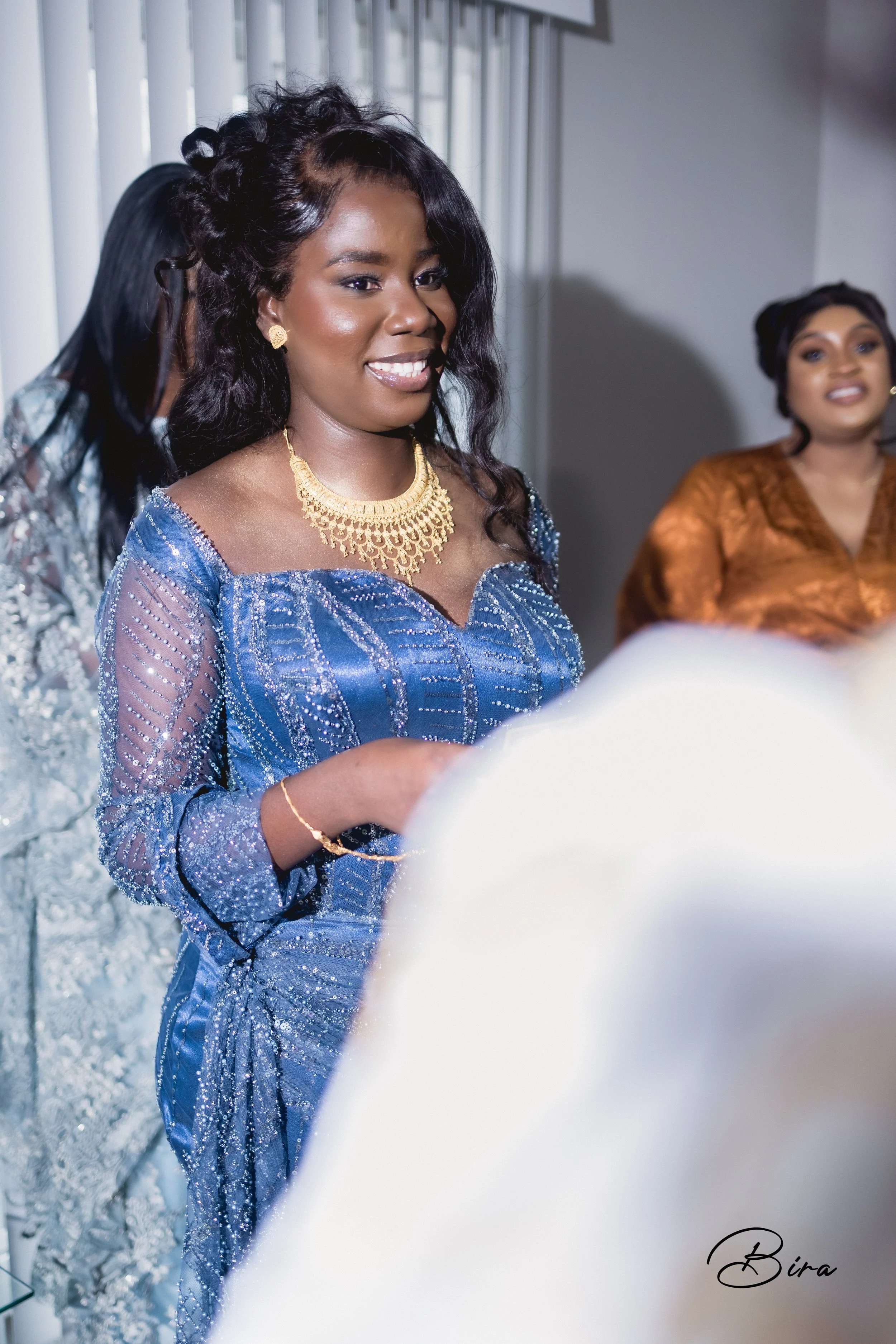 Two women at a formal event, one woman in the foreground wearing a sparkly blue dress and gold jewelry, smiling, with two other women in the background.