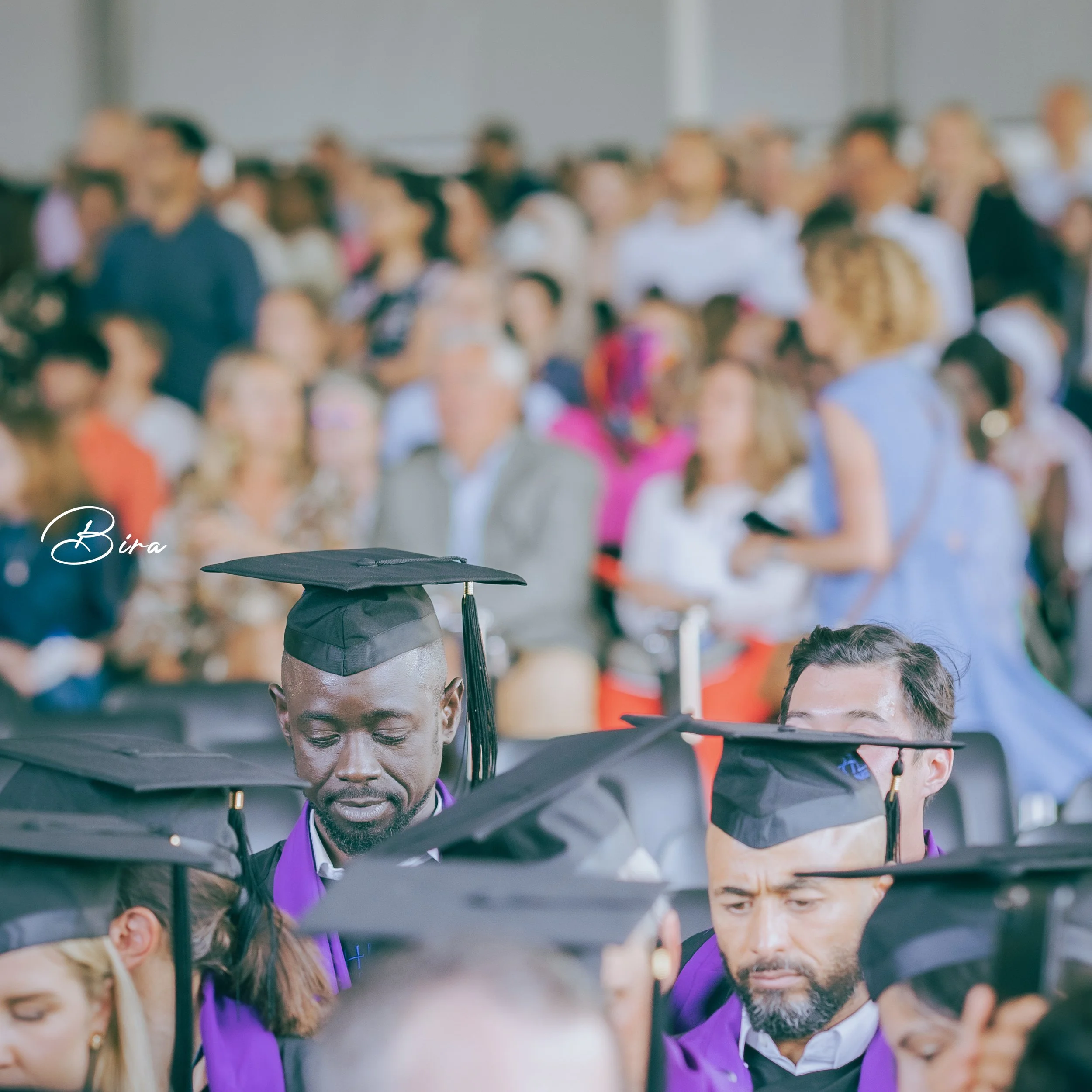 Graduates wearing caps and gowns at a ceremony, with a large crowd in the background.