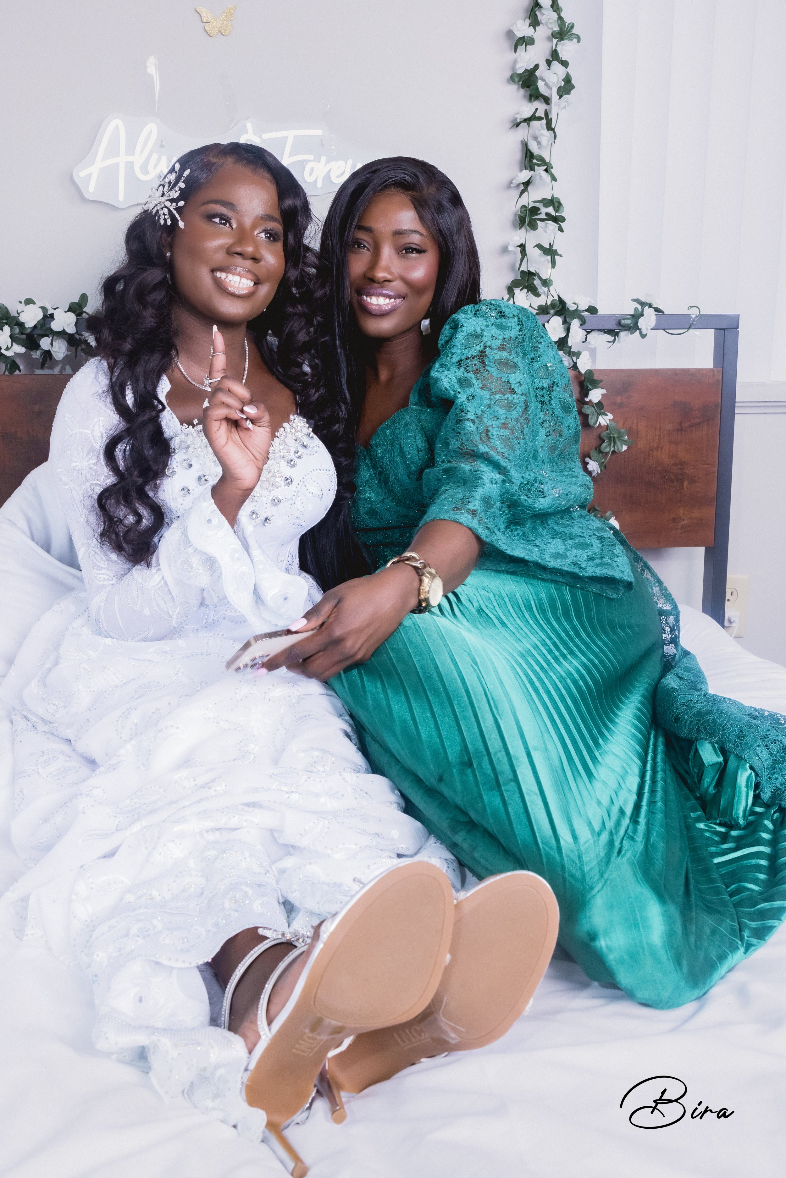 Two women sitting on a bed; one in a white dress with long wavy hair and the other in a green dress with black hair. They are smiling and making a peace sign. The background has a neon sign and floral decorations.