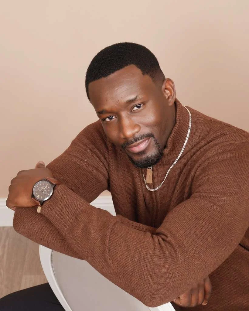 Portrait of a man with dark hair and beard wearing a brown sweater, a silver necklace, and a watch, sitting with his arms crossed on a white chair against a beige background.