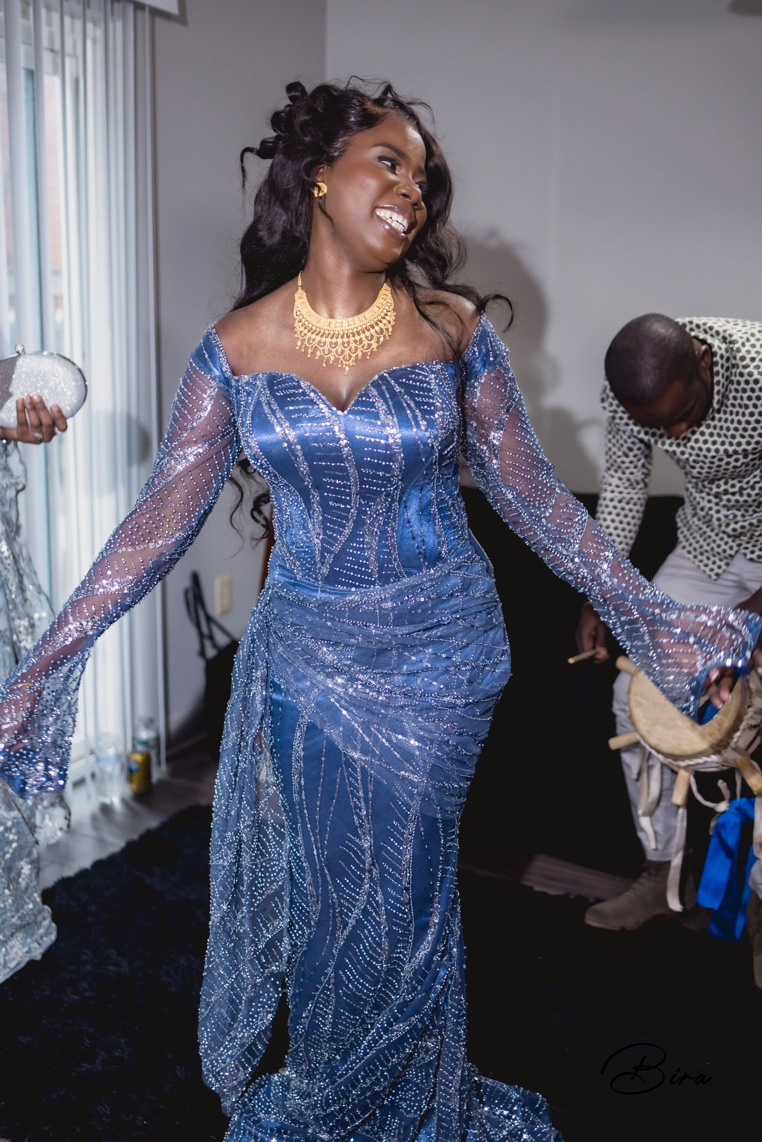 A woman in a sparkling blue dress with sheer sleeves and intricate beadwork, wearing gold jewelry, smiling and dancing in a room with a man in the background.