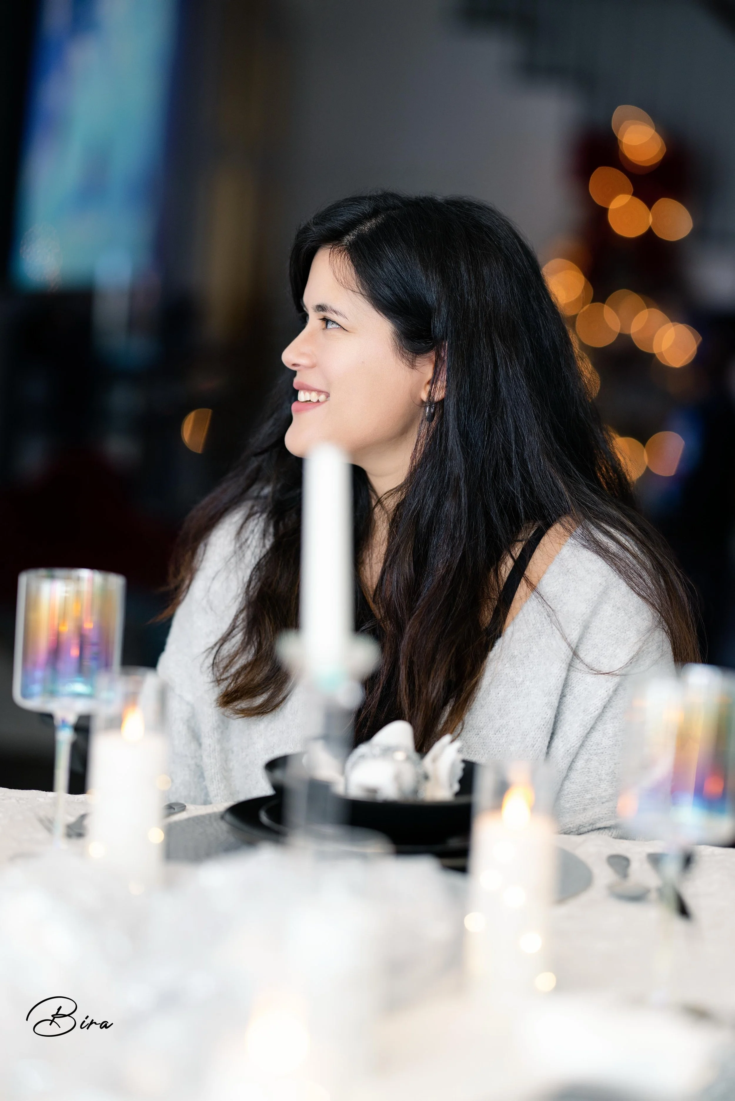 A woman with long dark hair is smiling and looking to the side, sitting at a table with lit candles and decorative glasses, in a dimly lit setting with blurred warm lights in the background.