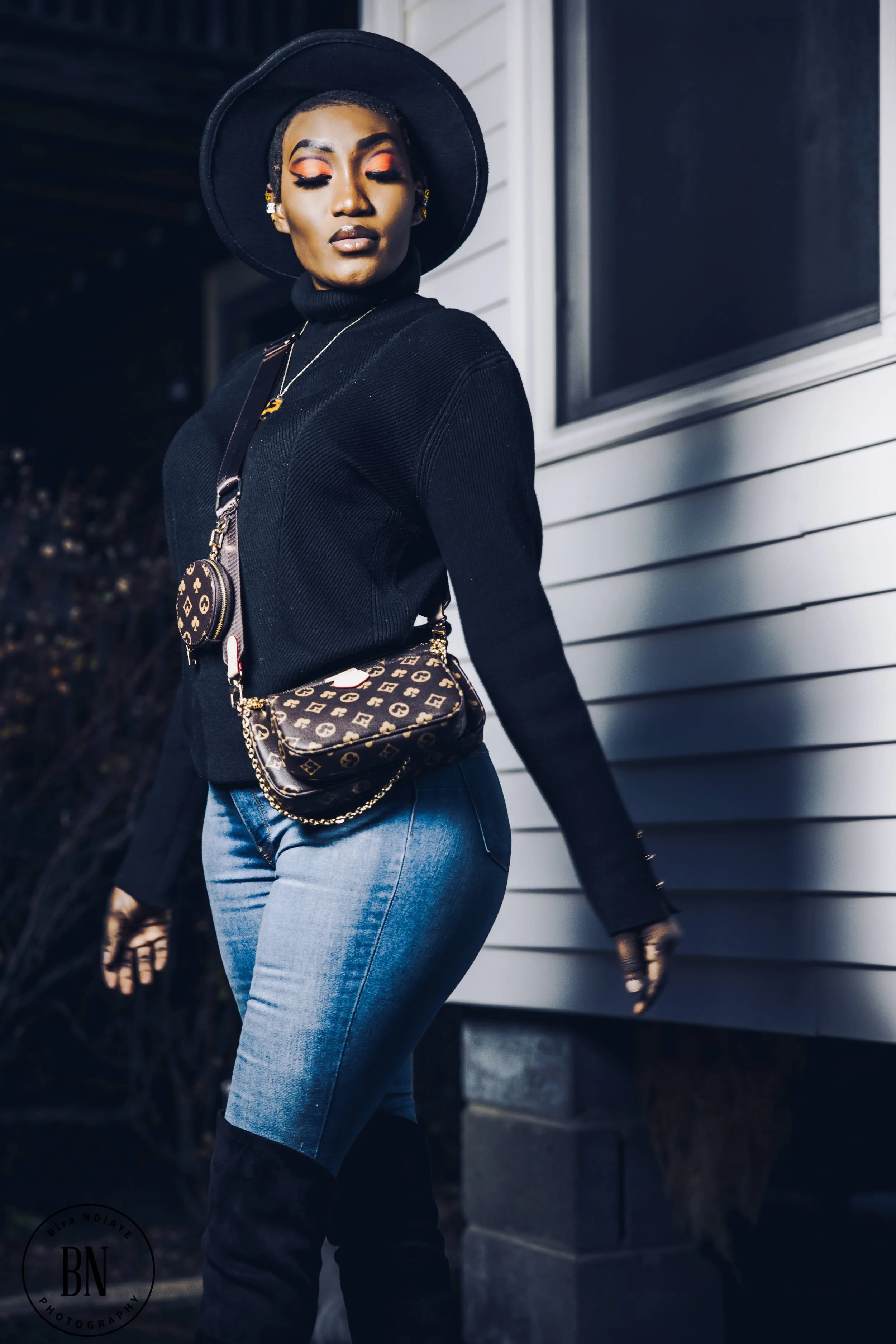 A woman dressed in black and blue denim standing outside at night, wearing a black wide-brimmed hat, black turtleneck sweater, and accessorized with a patterned crossbody bag, matching circular bag, earrings, and a necklace. She has her eyes closed a