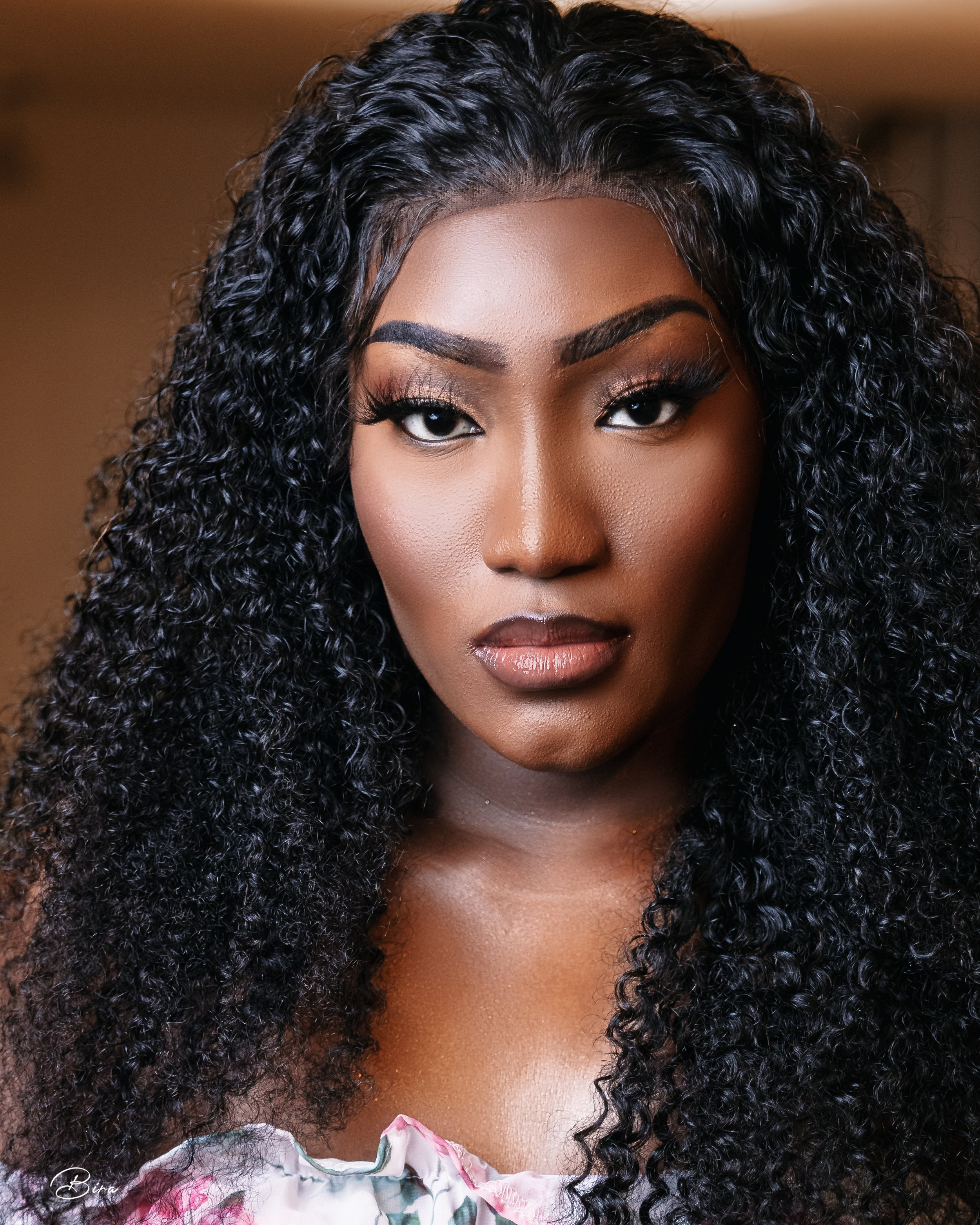 Close-up portrait of a woman with dark skin, curly black hair, and makeup, looking directly at the camera.