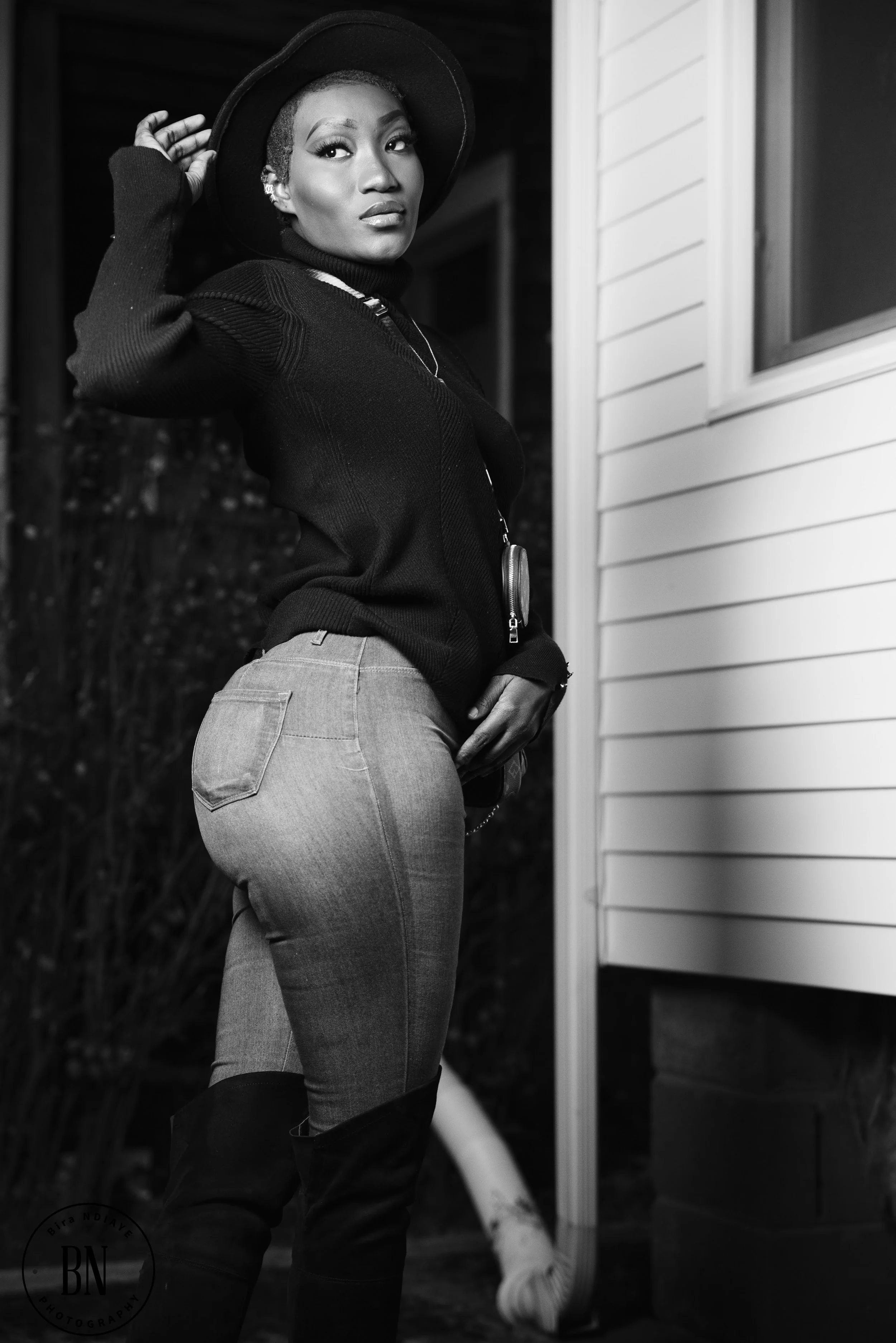A woman standing near a house in black and white, wearing a wide-brim hat, dark sweater, and jeans, with her hand on her hip and looking at the camera.