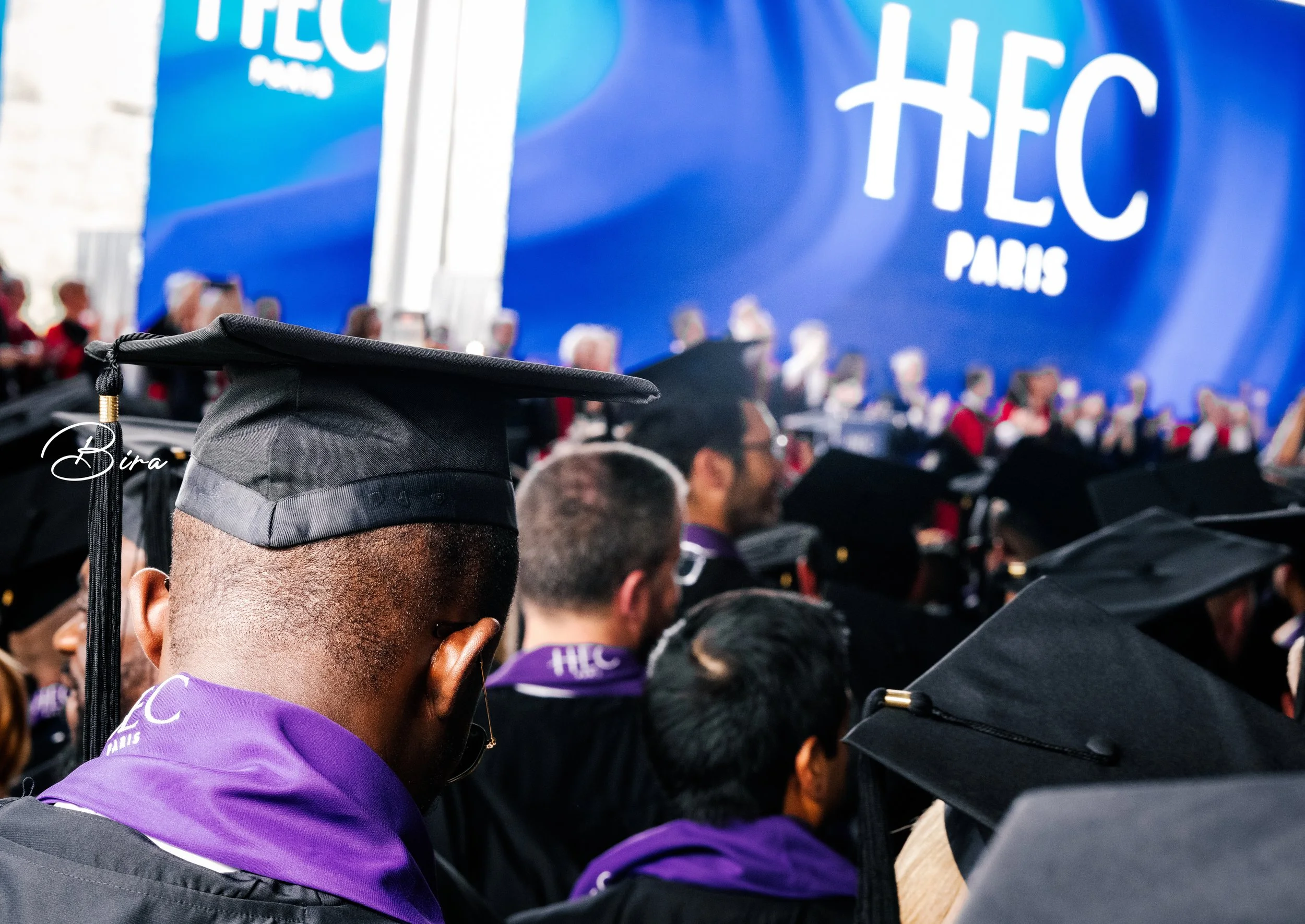 Graduates in black caps and gowns, some wearing purple scarves, attending a graduation ceremony at TEC Paris. A large blue TEC Paris banner is in the background.