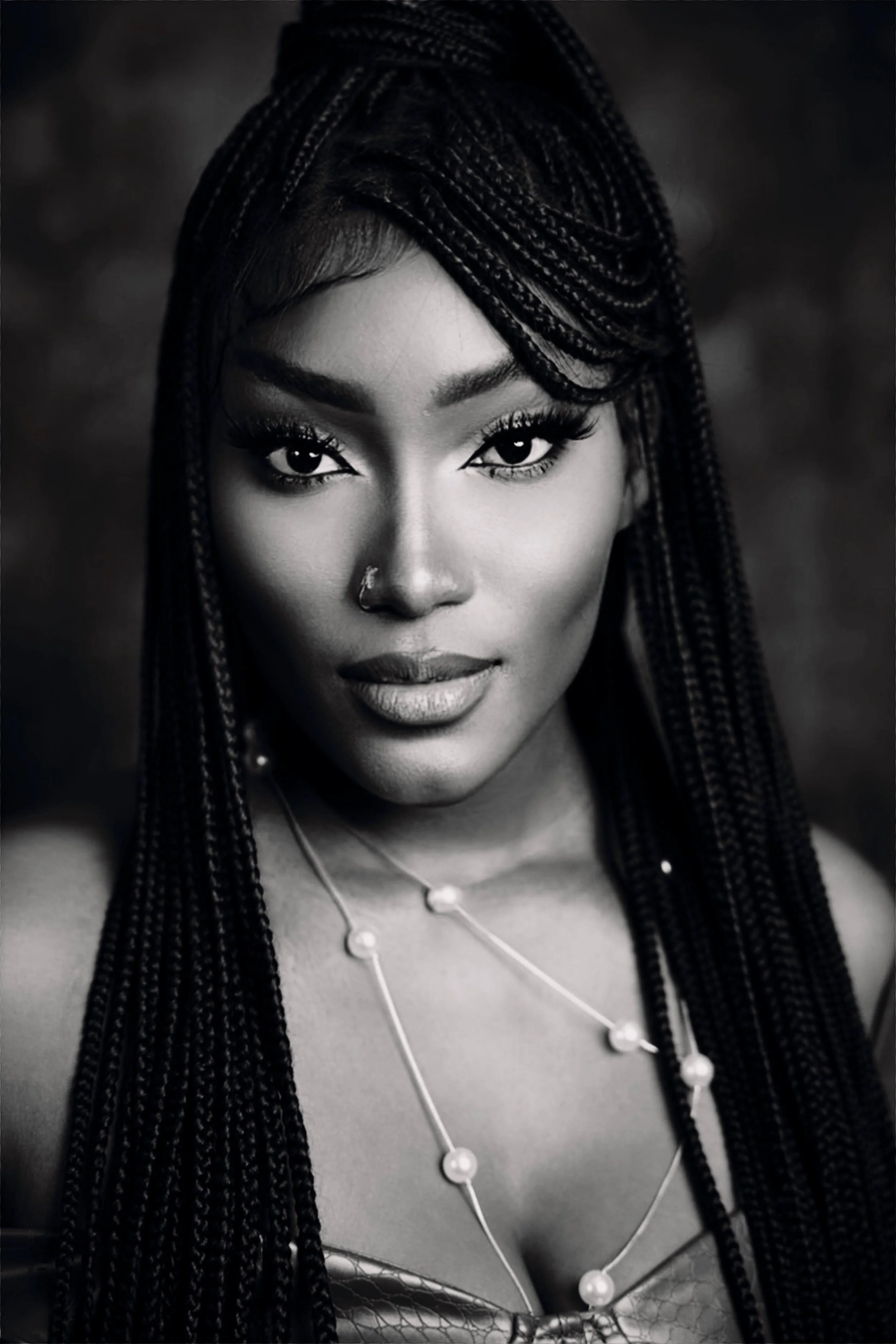 A black and white portrait of a woman with braided hair, wearing jewelry including a nose ring and a necklace with large pearls, looking directly at the camera.