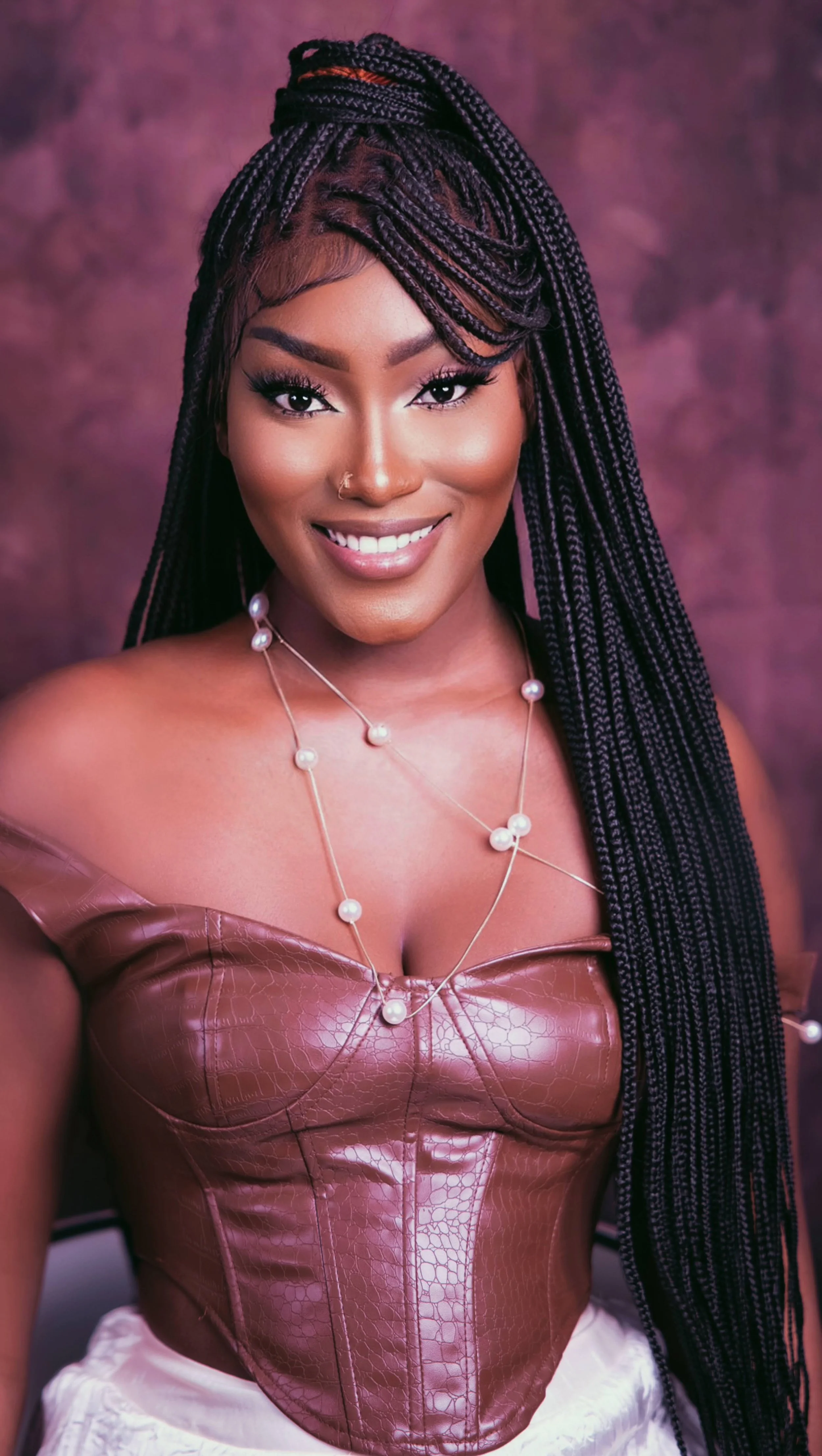 A young woman with long braided hair, smiling, wearing a pink off-shoulder top with a pearl necklace, against a pinkish-purple background.