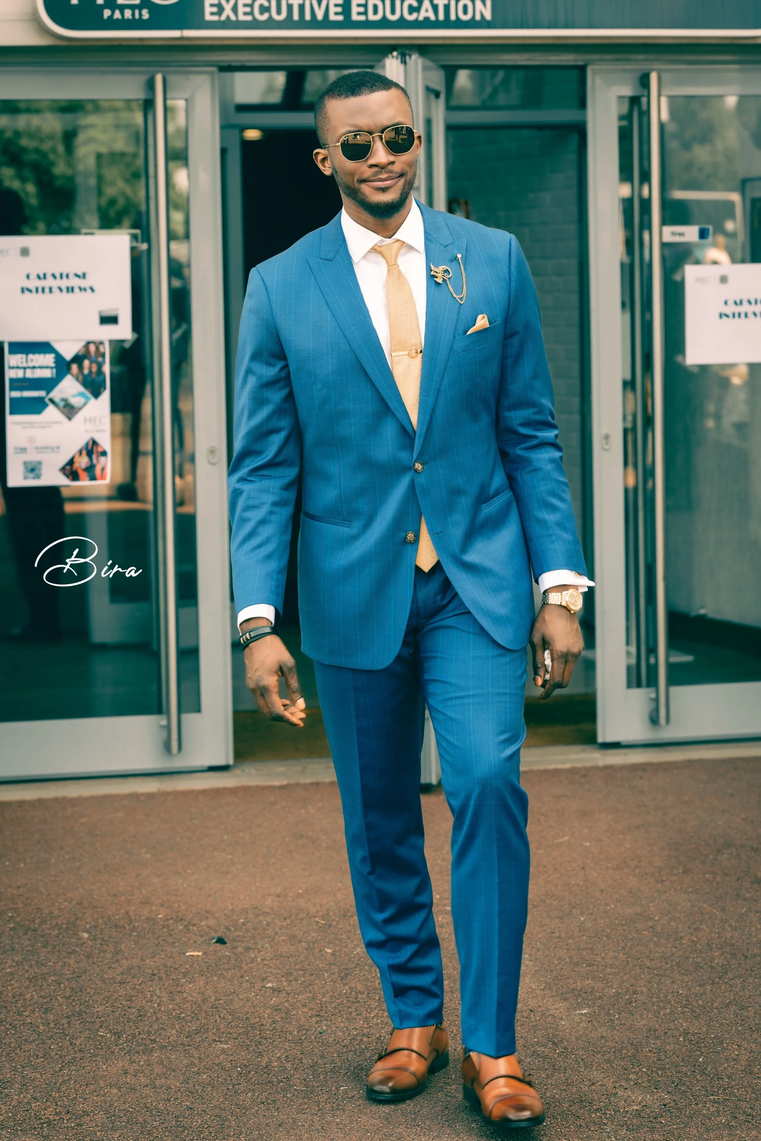 A man dressed in a stylish blue suit, white shirt, tan tie, and brown shoes, walking out of a building with glass doors. He is wearing sunglasses, a watch, and a gold lapel pin.