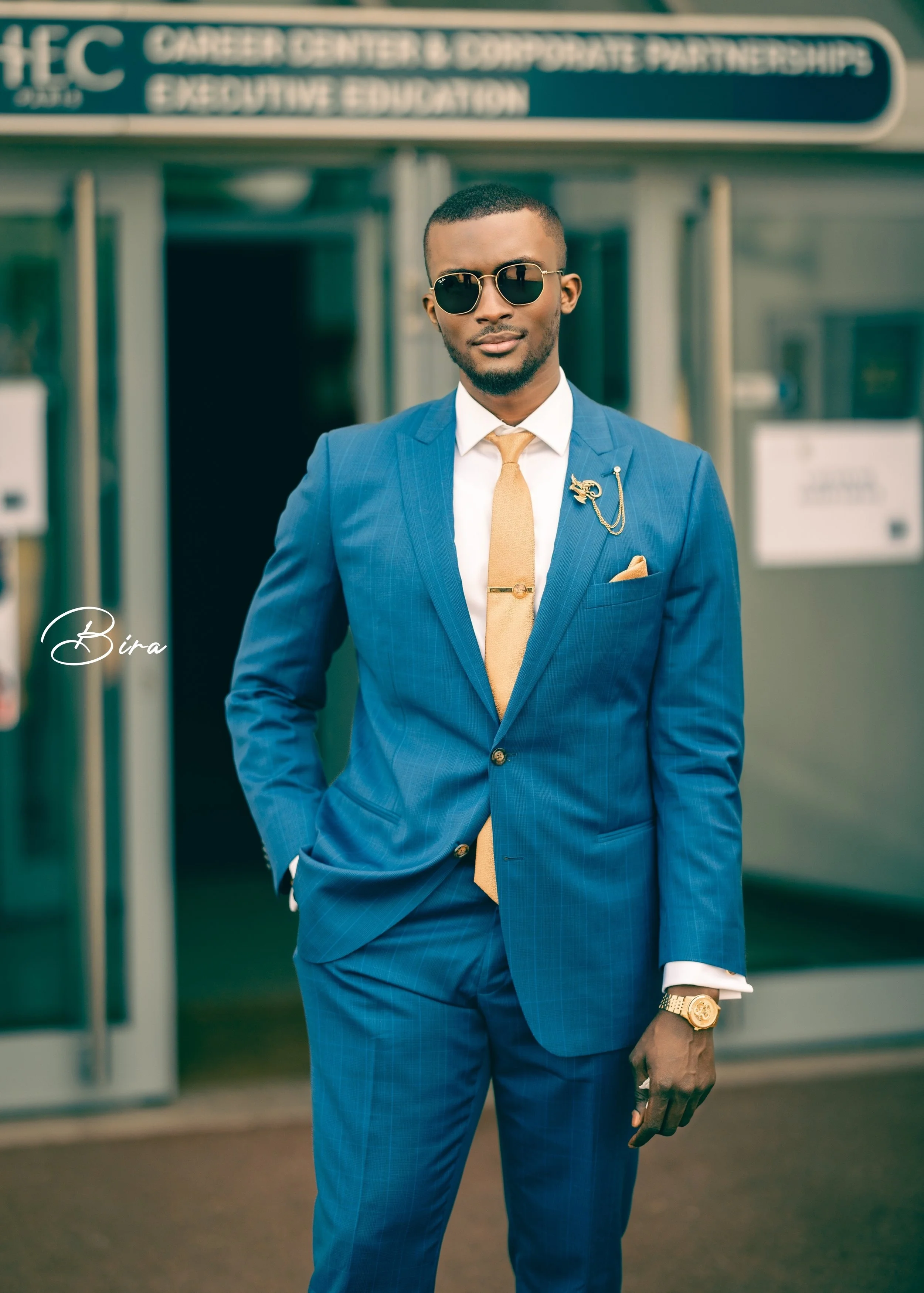 A young man in a blue suit with a white shirt and camel-colored tie, standing confidently outside a building with a career center sign, wearing sunglasses, a gold watch, and a decorative lapel pin.