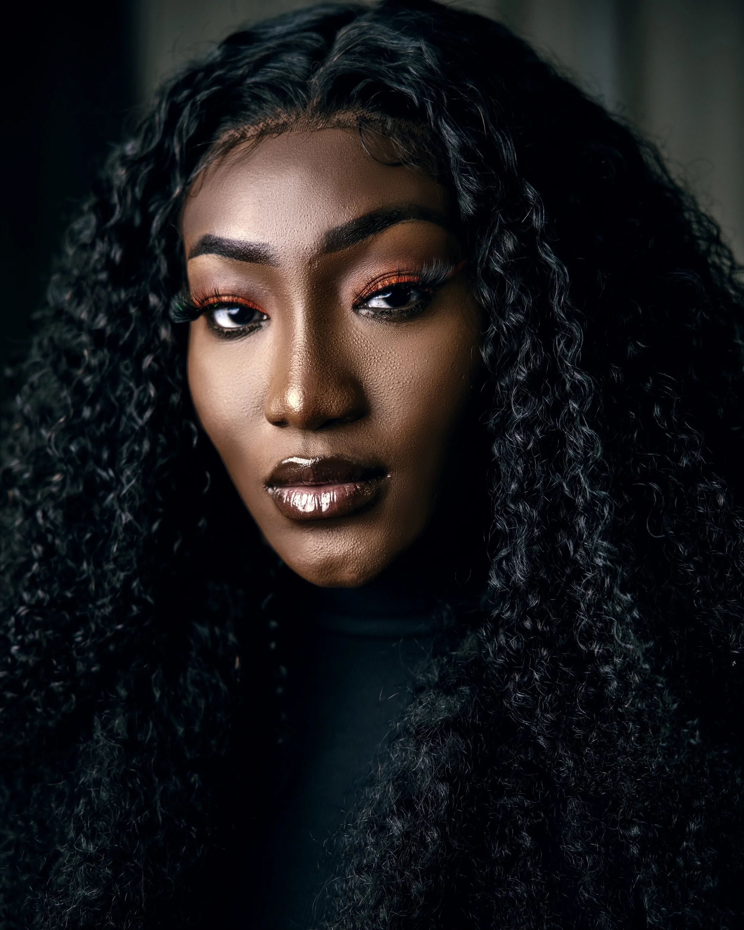 Close-up of an African American woman with curly hair, wearing makeup with black eyeliner, orange and gold eyeshadow, and glossy dark lipstick