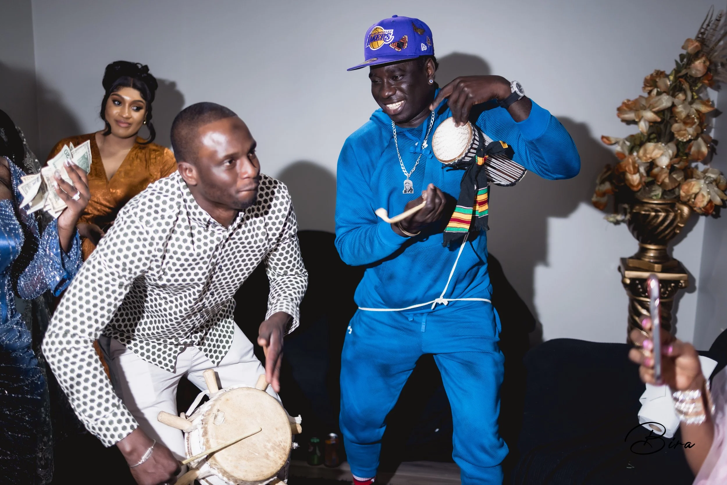 People celebrating at a gathering, with some playing drums and others dancing, in a room with flowers in a vase.