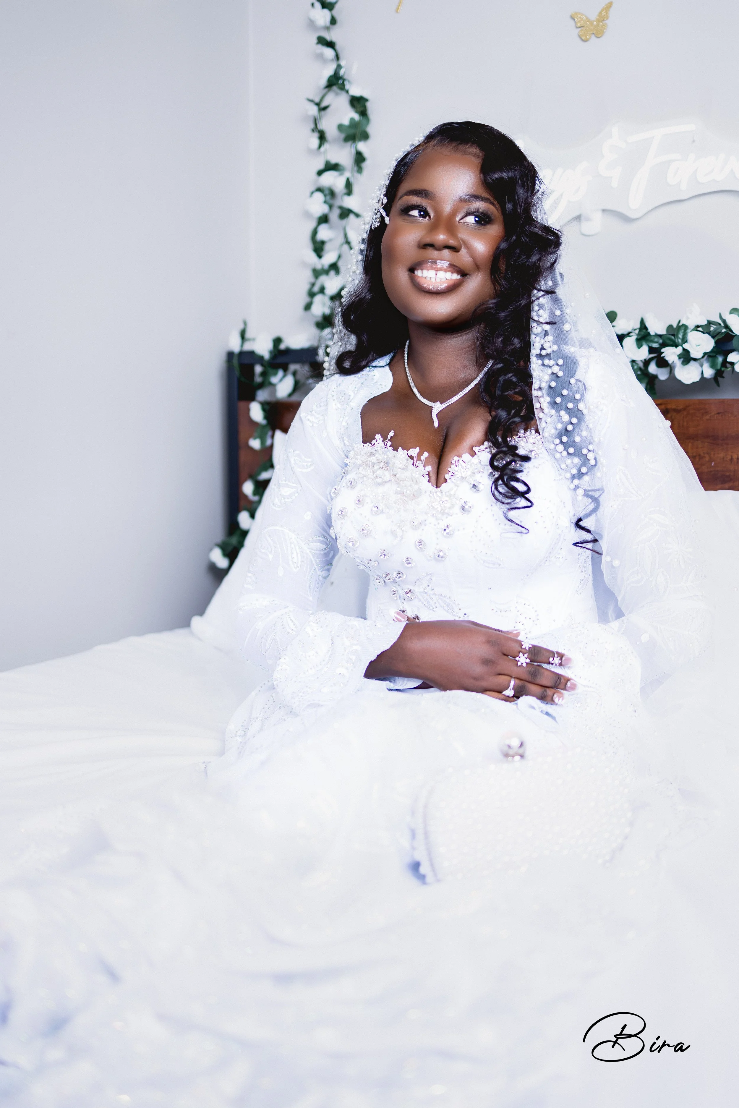 A smiling bride with dark curly hair in a white wedding dress, sitting on a bed in a decorated room with white and green accents.